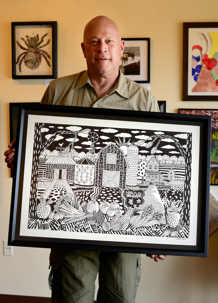 An indoor photograph of Fred Seger holding up his framed black and white ink-based artwork to the camera.