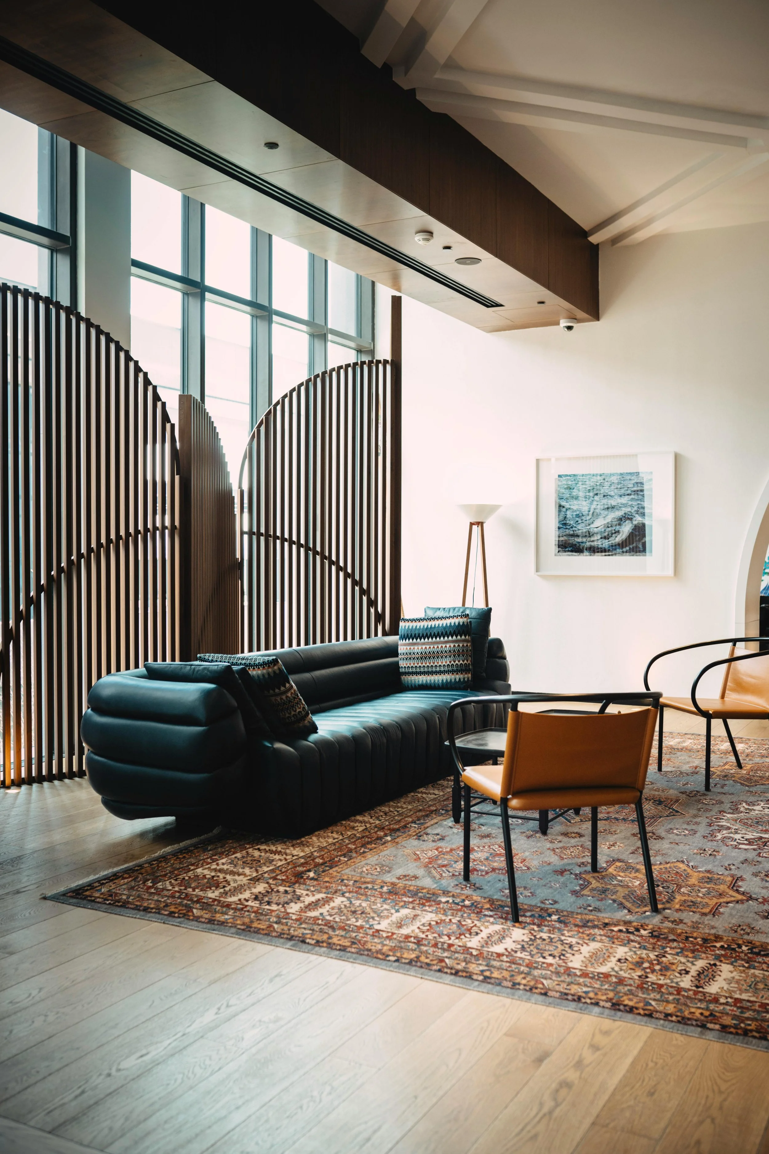 Interior of a modern lounge or waiting area with large windows, a black leather sofa, a rug, a framed artwork, and chairs, with wooden wall panels and natural light.