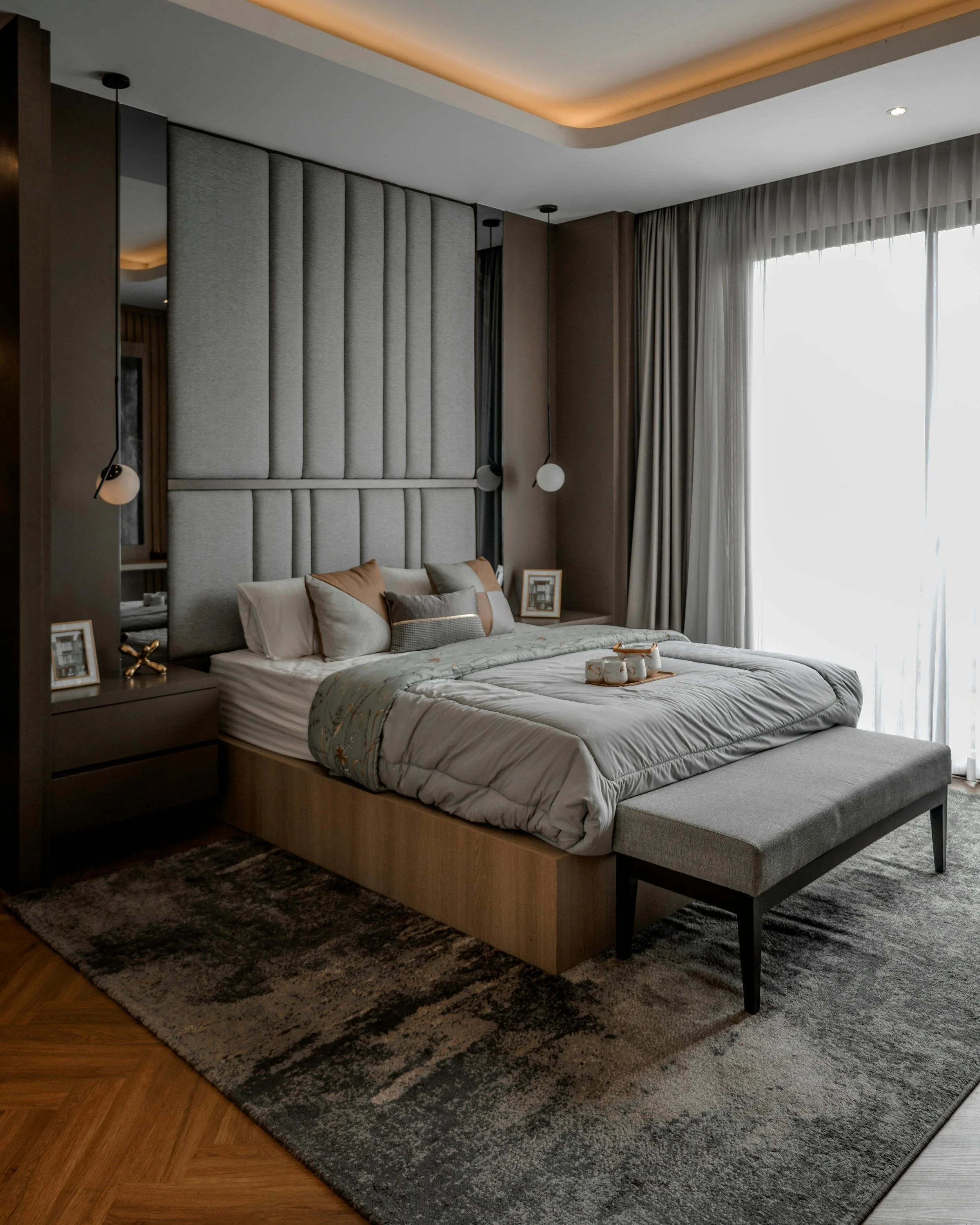 A modern bedroom with a large bed, cushioned headboard, throw pillows, and a tray with cups on the bed. The room features a window with sheer curtains, a bedside table with framed photos, and pendant lights hanging from the ceiling.