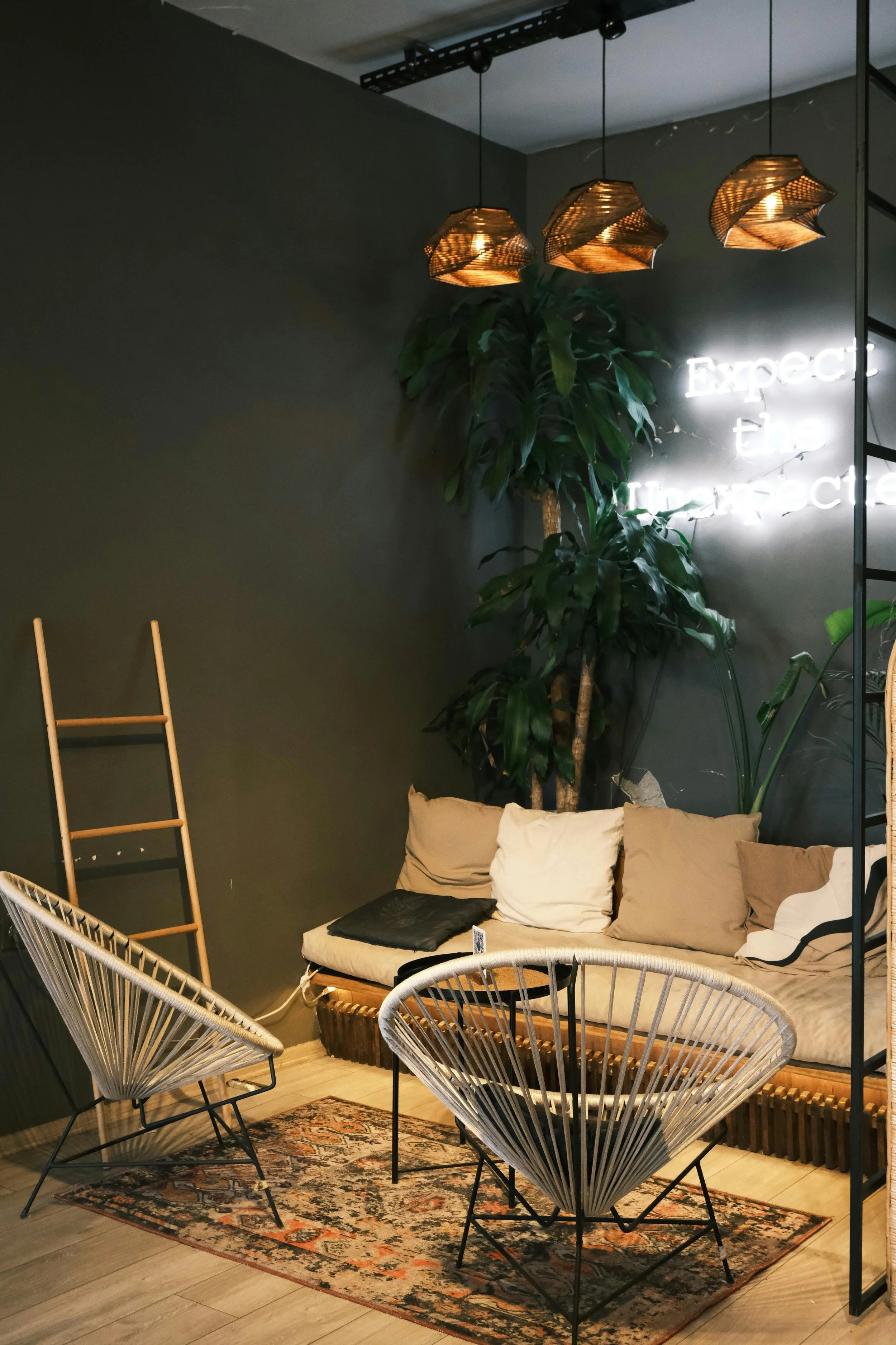 A cozy indoor seating area with a patterned rug, two wire armchairs, a wooden outdoor-style sofa with beige and white cushions, a large green indoor plant, a wall with a neon sign, three hanging woven pendant lights, and other decorative elements.