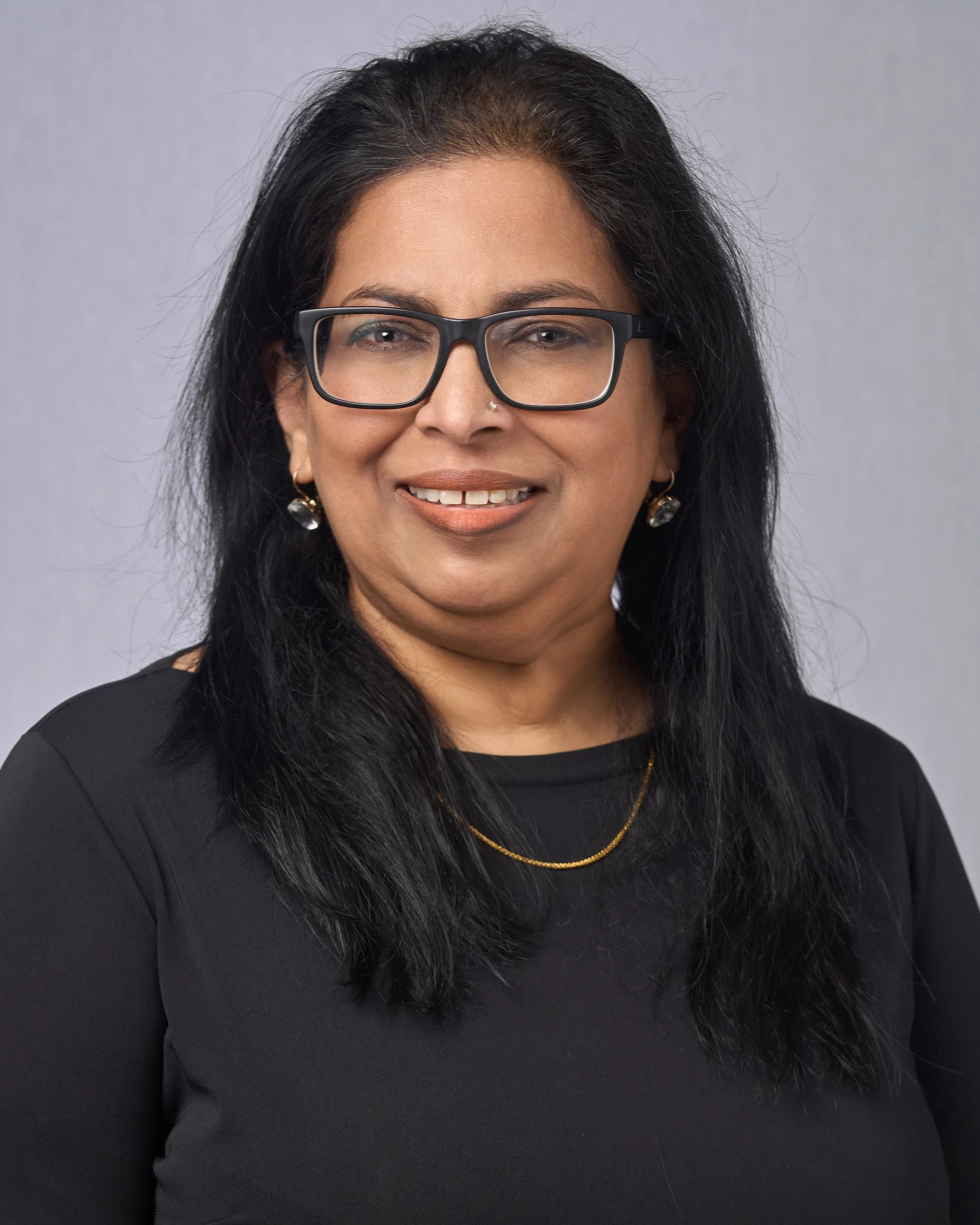 Woman with black hair, glasses, hoop earrings, and a gold necklace, smiling, wearing a black top, against a neutral background.