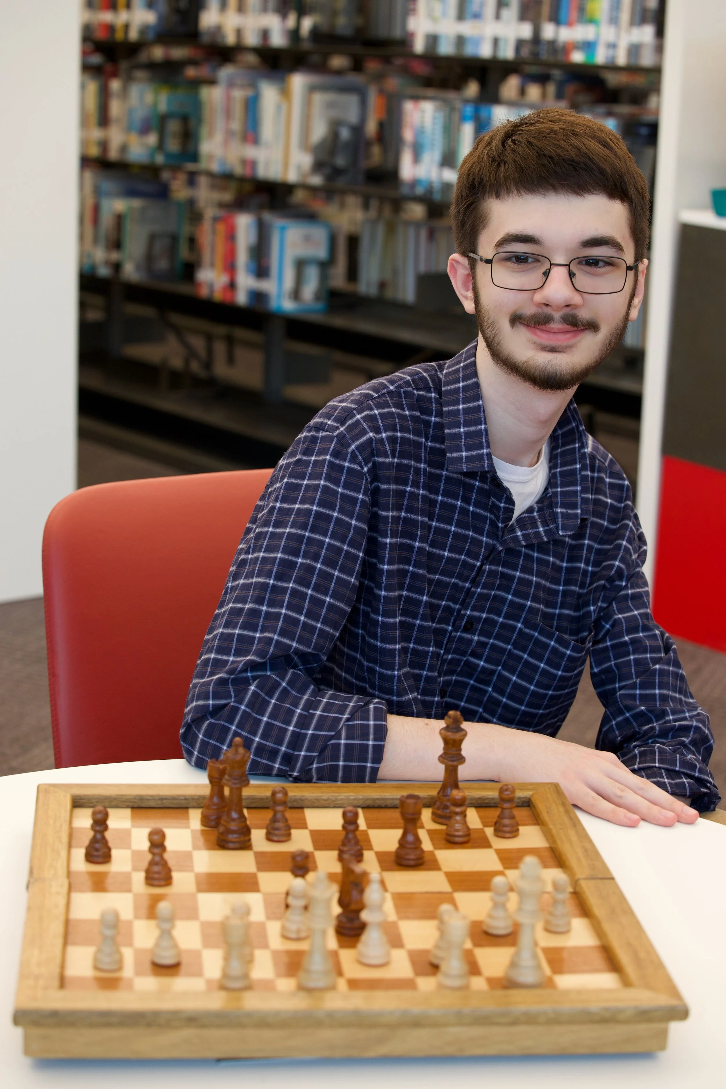 A young man with glasses and a beard, wearing a blue checkered shirt, sitting at a table with a chessboard, smiling. The background includes bookshelves in a library.
