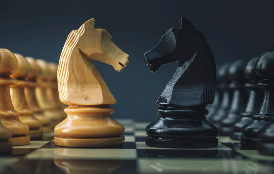 Close-up of a wooden light-colored chess knight facing a black chess knight on a chessboard.