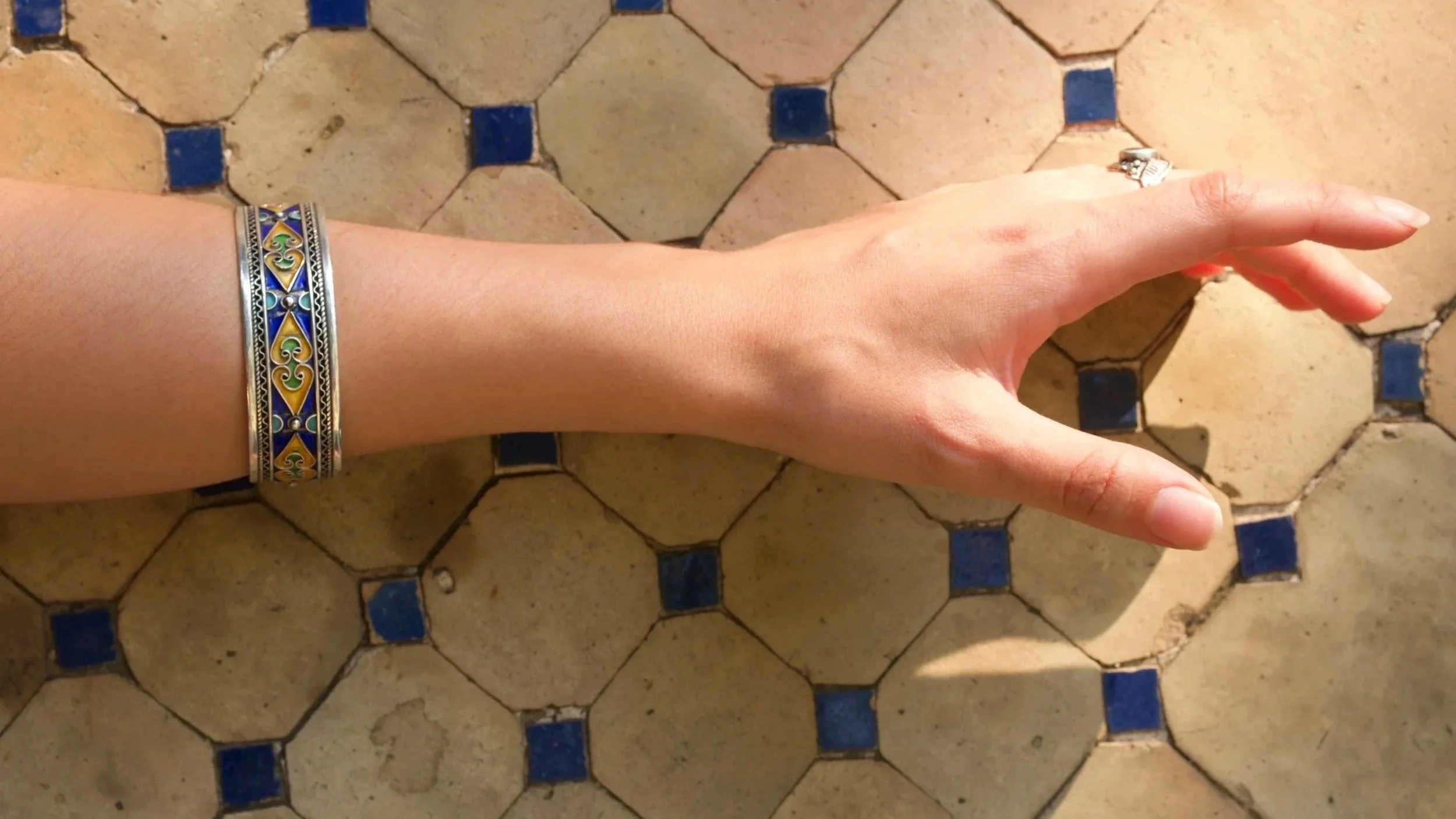 A person's arm and hand extended over a vintage tiled floor. The arm is adorned with three colorful, patterned bracelets. The floor has an intricate geometric pattern with beige tiles and small blue square accents.