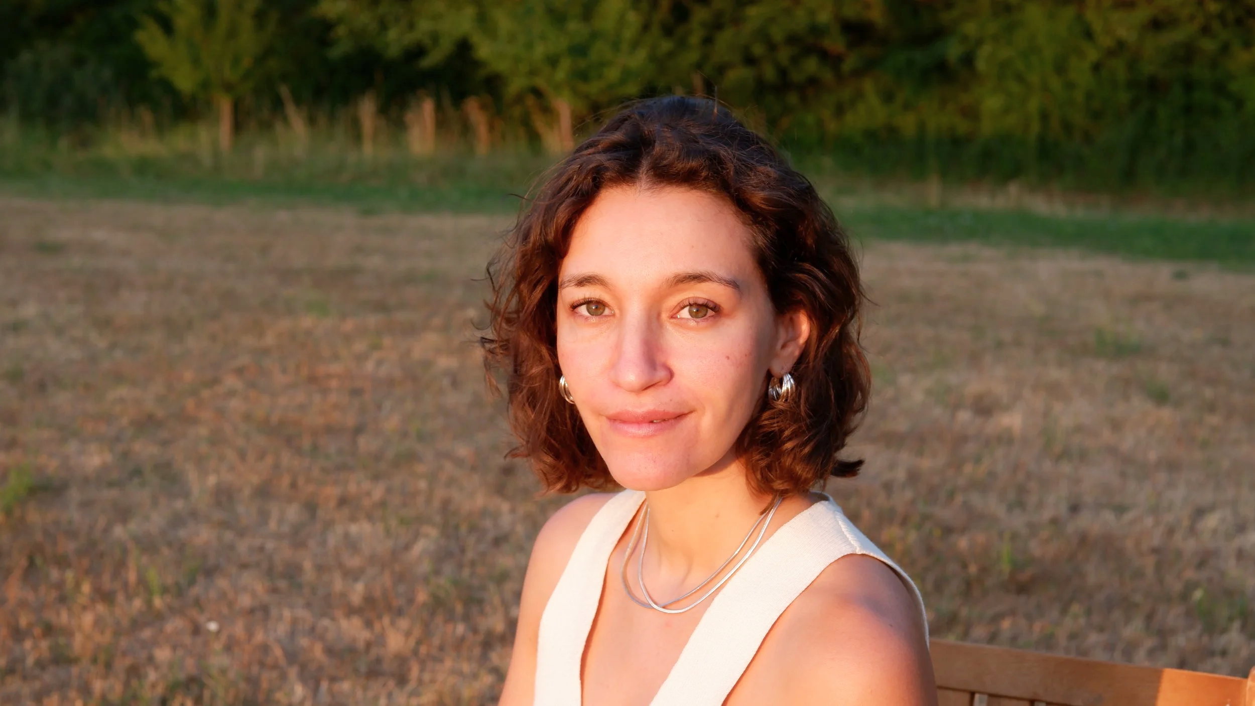 A woman with short, curly brown hair and light green eyes sitting outdoors during sunset, wearing a cream sleeveless top and silver jewelry, smiling softly at the camera with a natural landscape in the background.