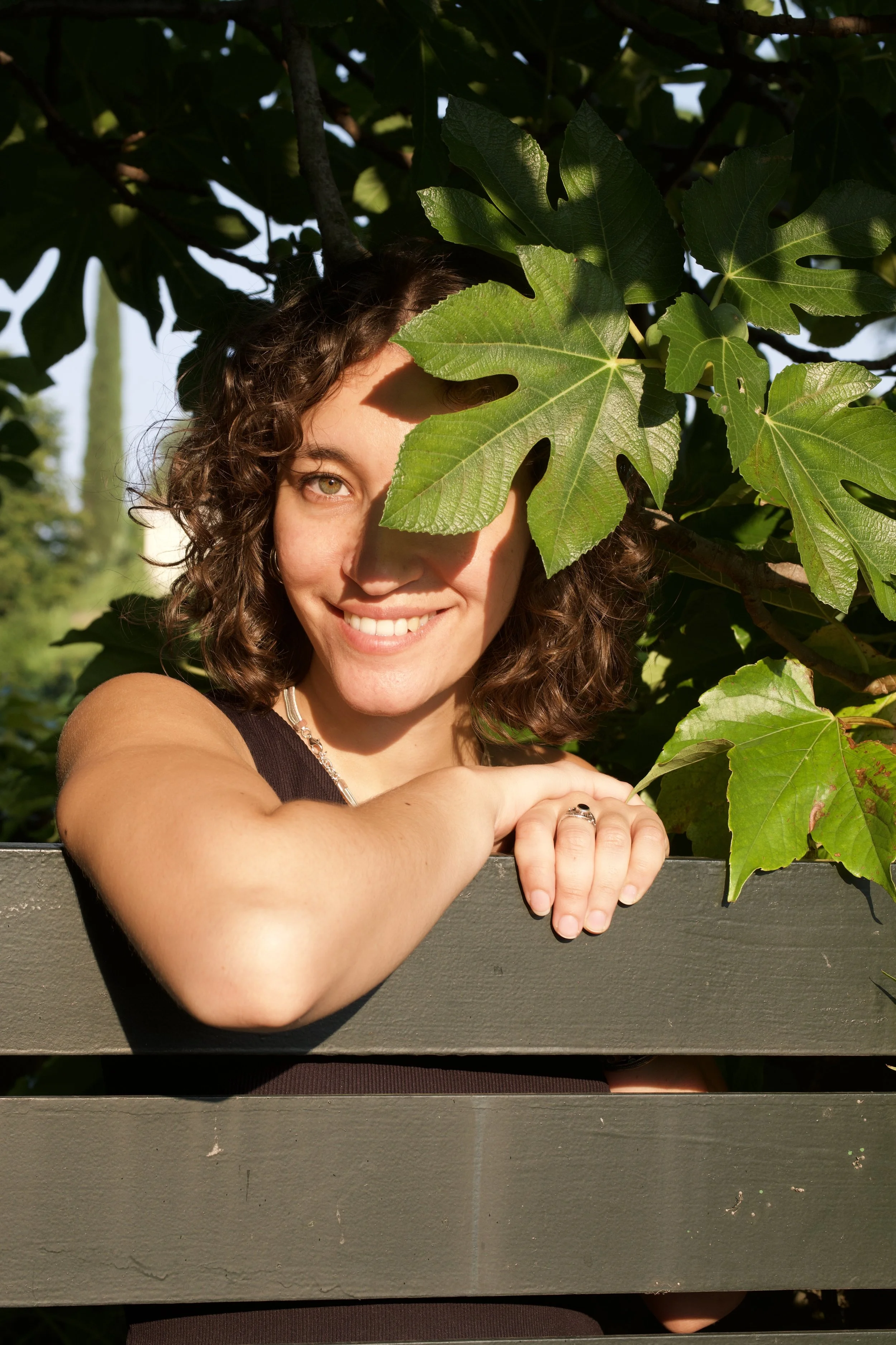 A woman with curly brown hair smiling, partially hidden behind large green leaves, leaning on a gray wooden ledge outdoors on a sunny day.