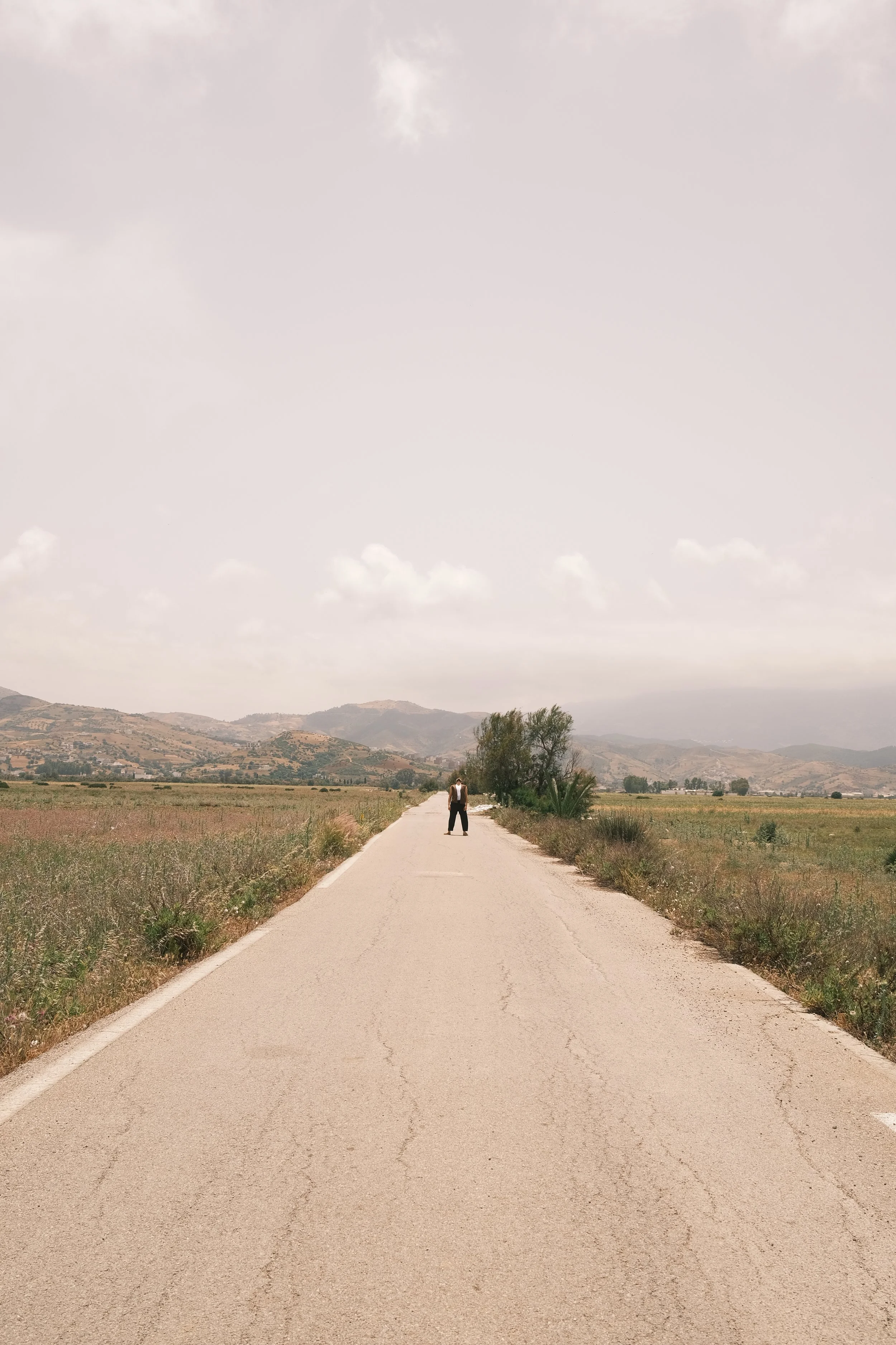 A person standing alone in the middle of an empty rural road in a vast open landscape with mountains in the distance and a cloudy sky overhead.
