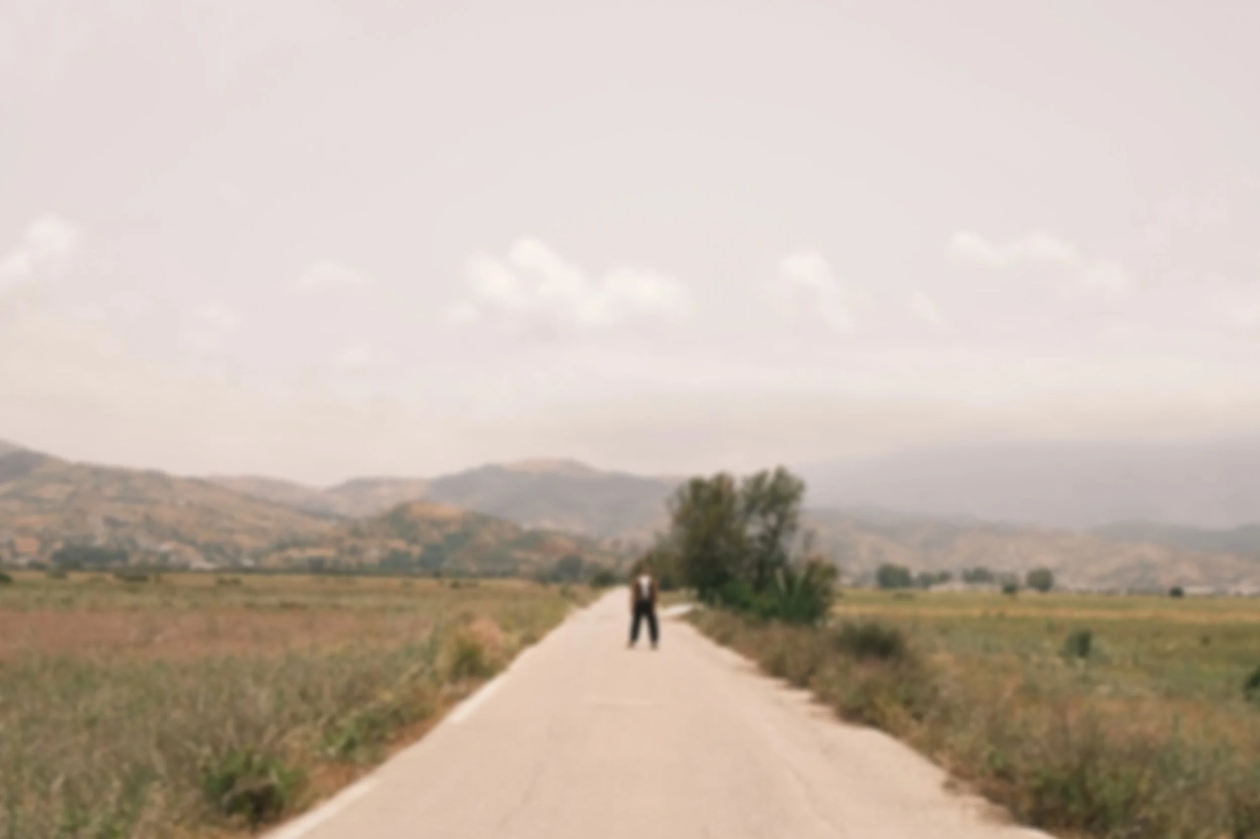 A person walking on a rural road through a landscape with fields, mountains in the background, and a partly cloudy sky.