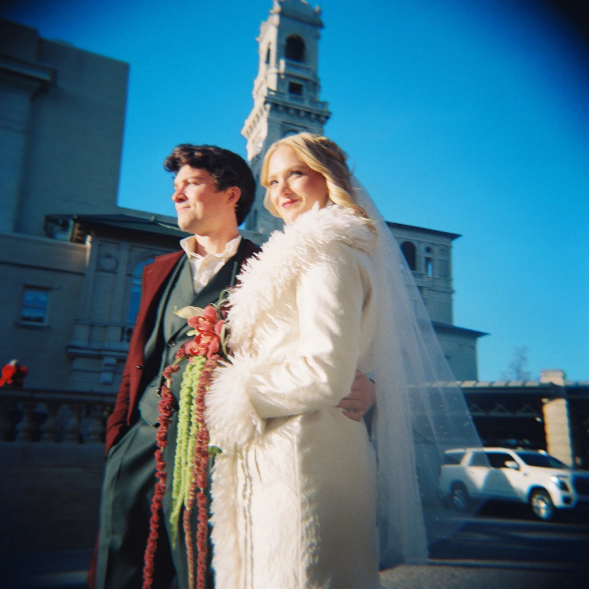 A man and woman dressed in wedding attire, standing outdoors in front of a tall building under a clear blue sky. The woman is wearing a white fur coat while holding a bouquet, while the man is in a suit and tie.
