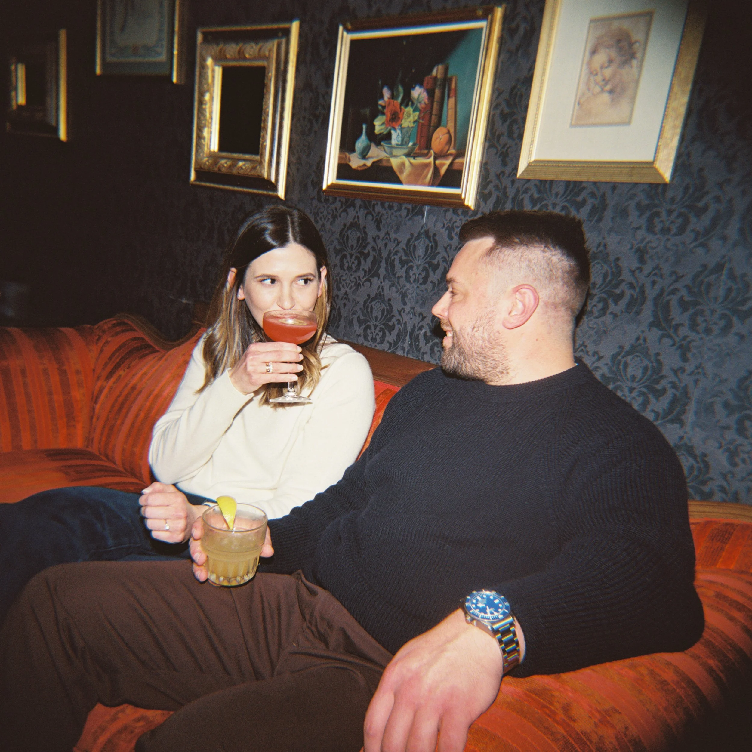 A woman with long brown hair and a man in a black shirt sitting together at a restaurant or bar, holding hands, with drinks in front of them. The woman is drinking a cocktail, and both are smiling. The background shows framed artwork and dark wallpaper.