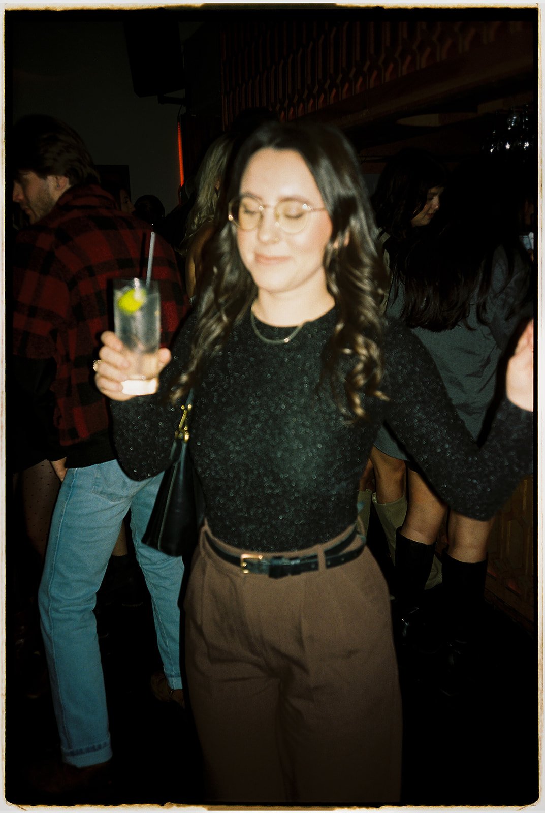 A woman with glasses and wavy hair holding a drink with a lime wedge and straw, dancing at a social gathering.