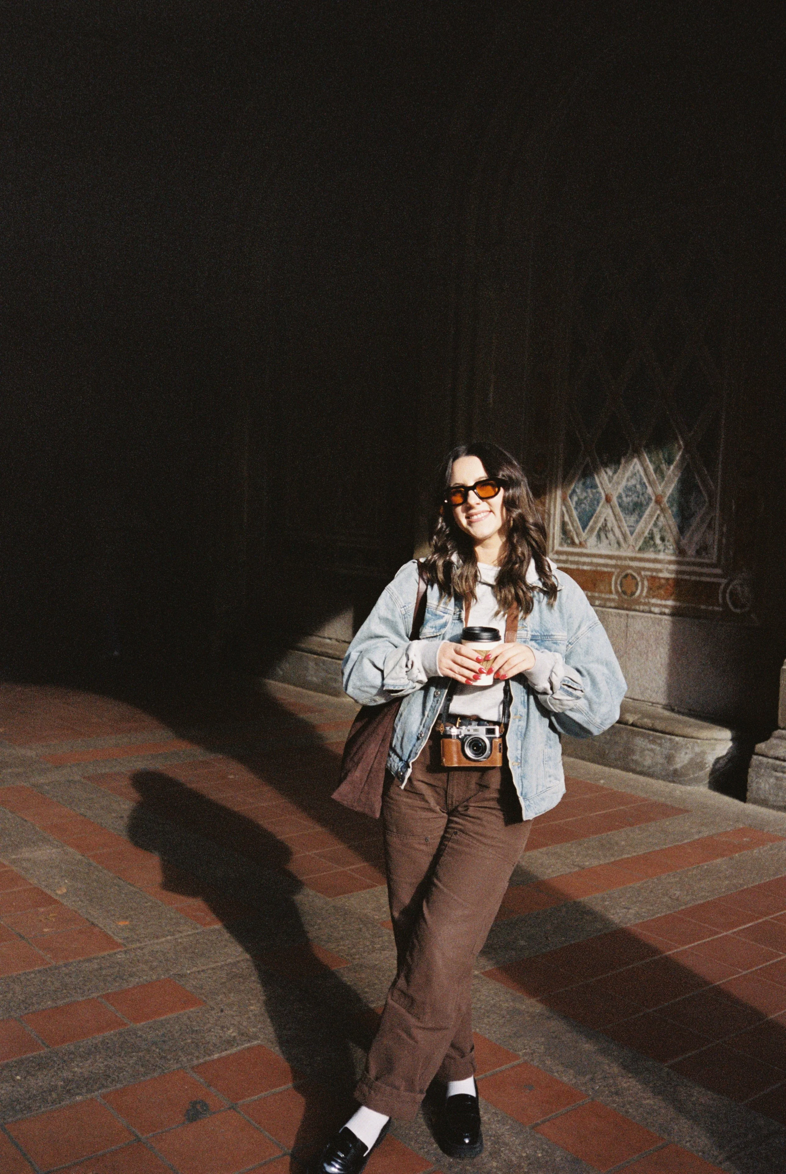 A young woman standing outdoors against a decorative wall, smiling, holding a coffee cup, wearing sunglasses, a denim jacket, brown pants, and black-and-white loafers.