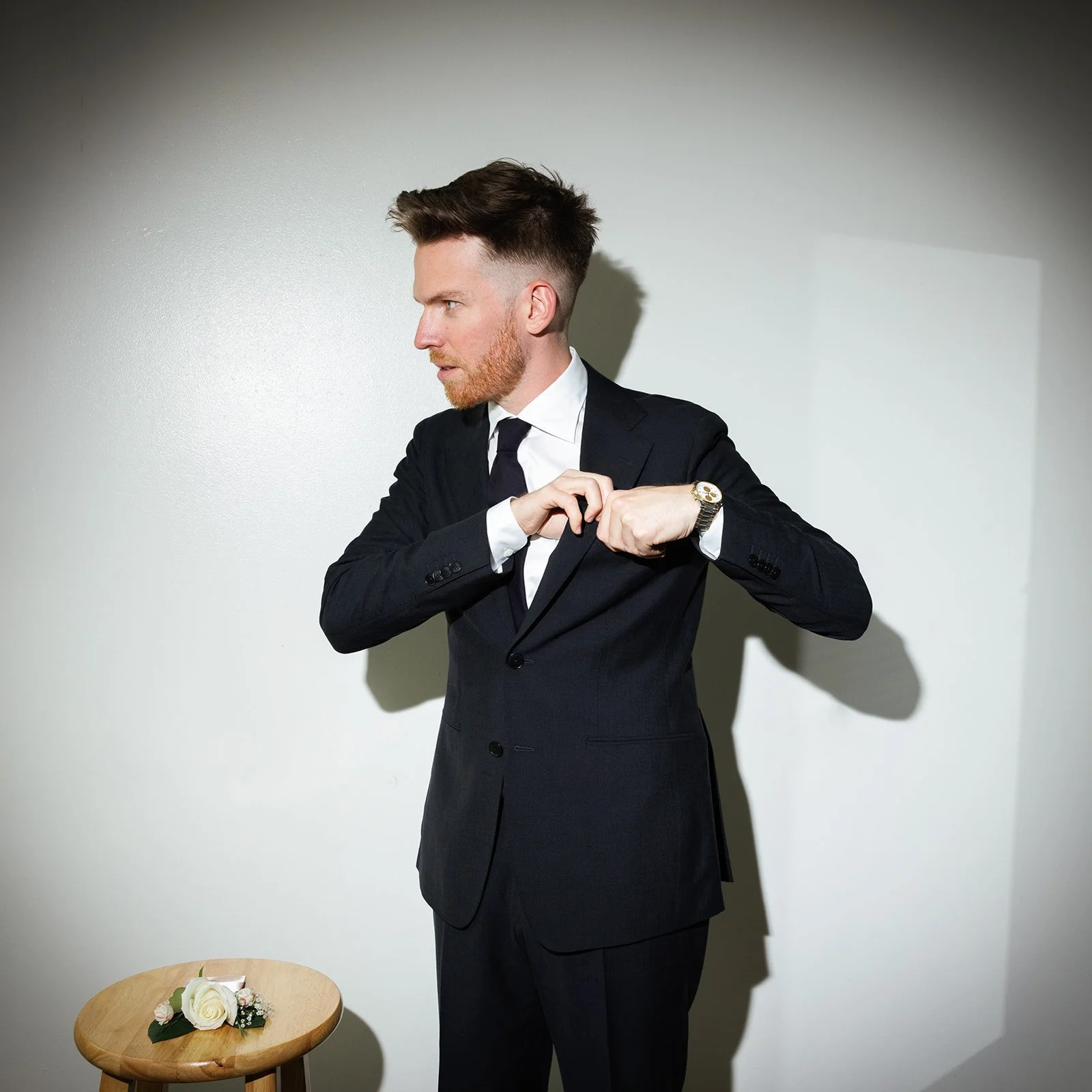 A man in a black suit and tie, with a white shirt, is adjusting his suit jacket against a plain white wall. A small round wooden table is in the foreground.
