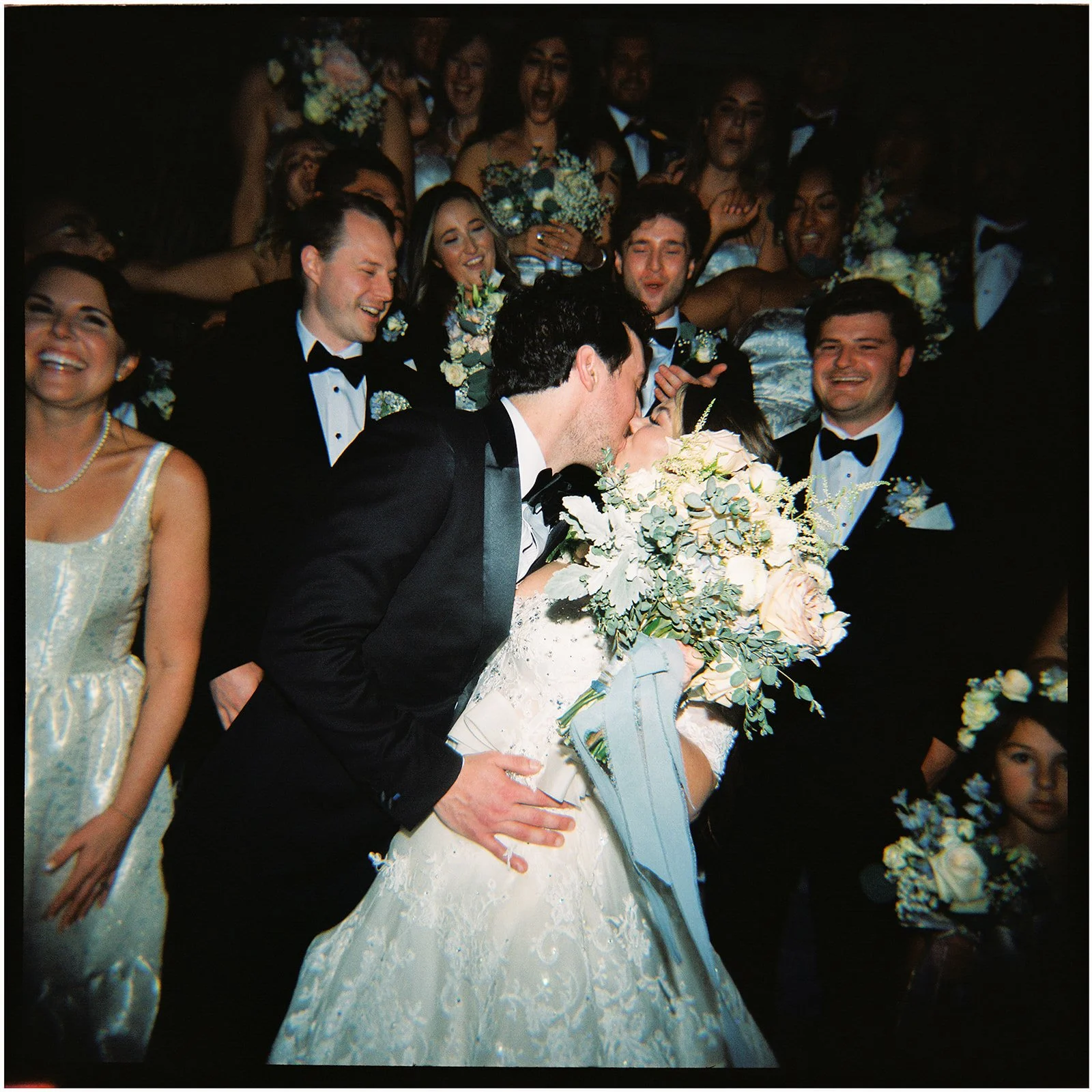 A film photo of a group of people at a wedding, with the bride and groom kissing in the center, surrounded by friends and family who are smiling and celebrating.