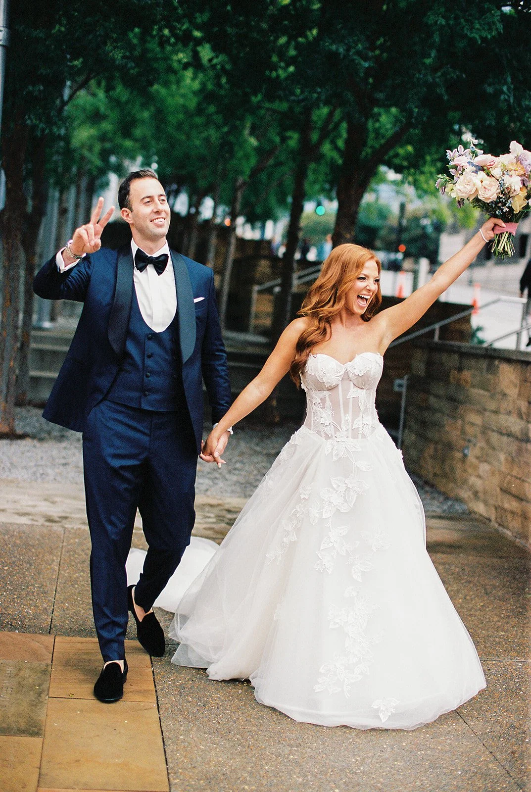 A bride and groom holding hands, celebrating outdoors. The bride, with red hair, is wearing a strapless white wedding gown with floral lace details and holding a bouquet of flowers and the groom, wearing a dark blue tuxedo with a black bow tie.