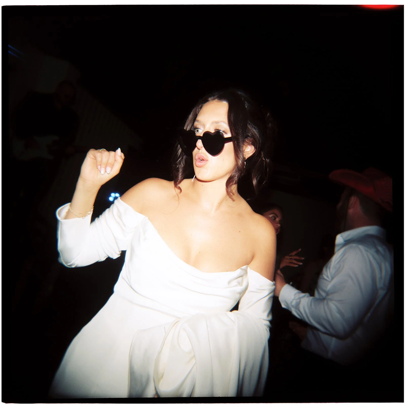 A woman dancing at a party wearing black heart-shaped sunglasses and a white off-the-shoulder dress.