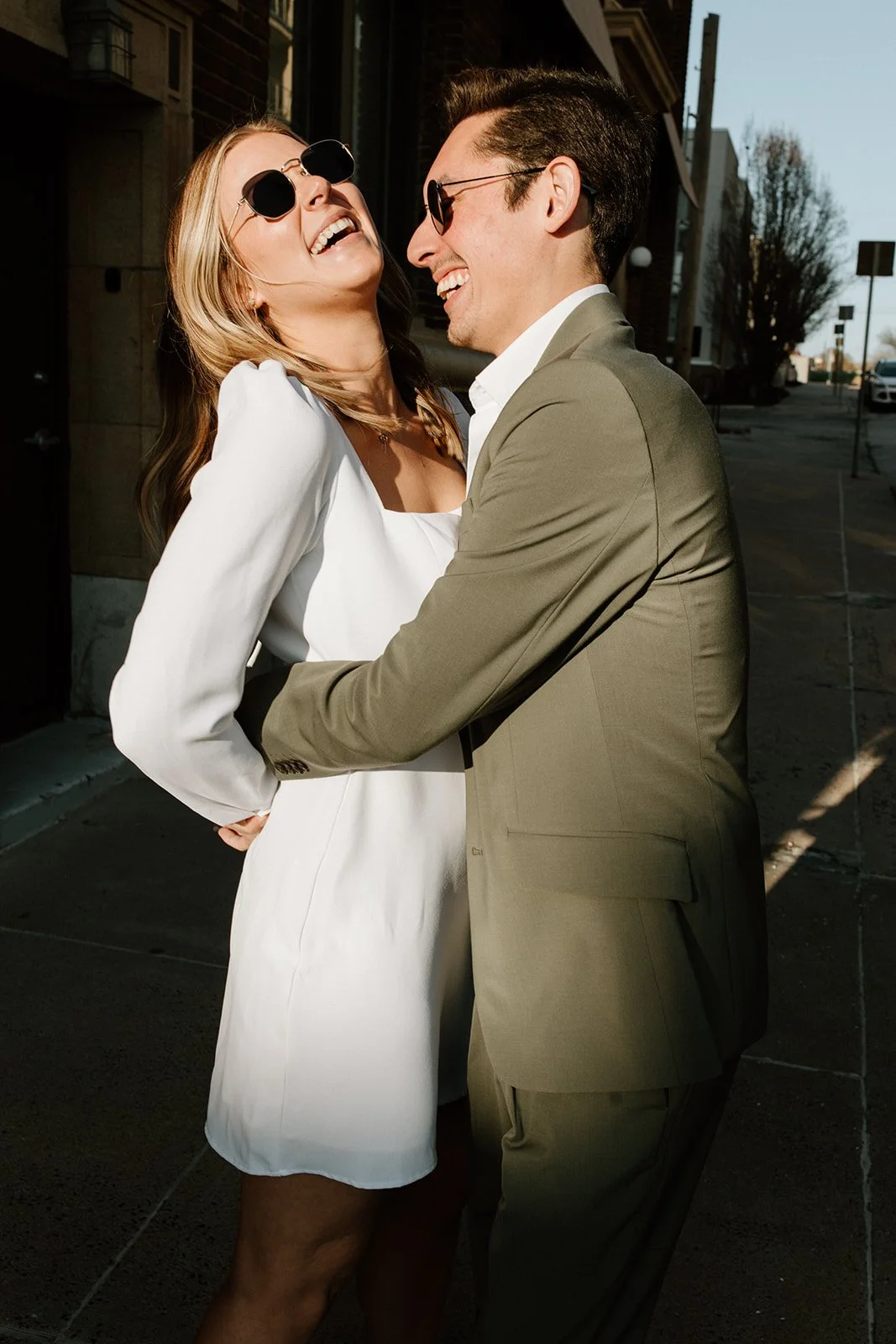 Woman wearing white dress smiling at man in green suit both with sunglasses on