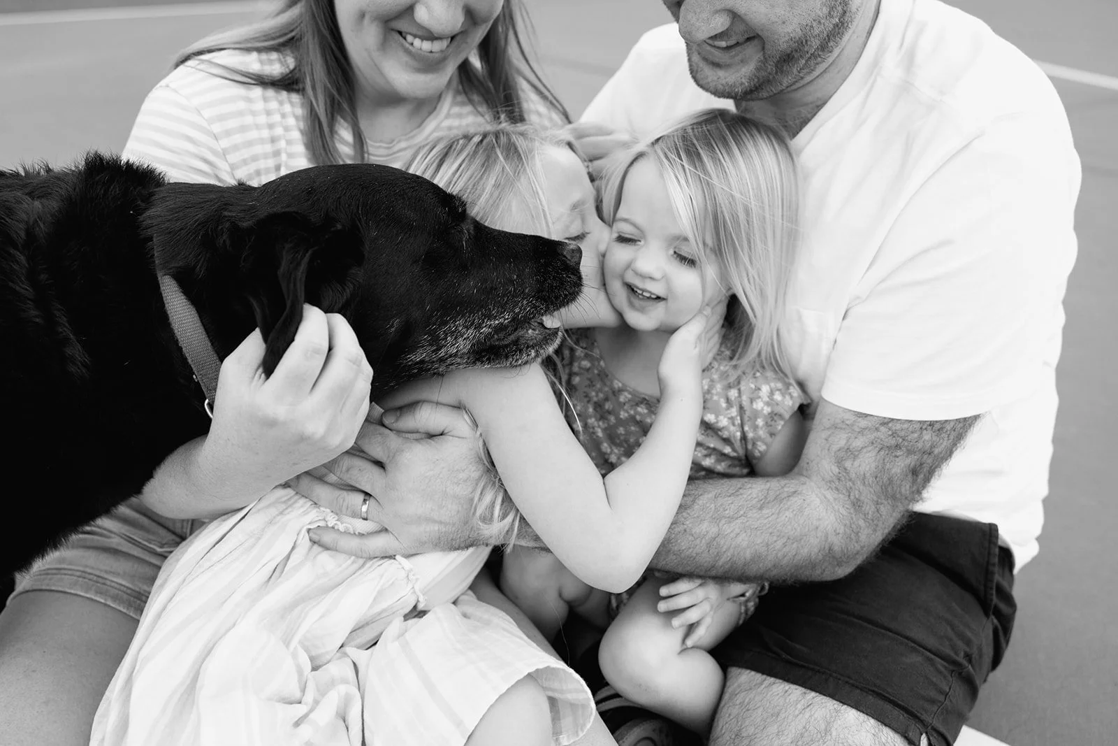 A happy family of four, including two young girls, a woman, and a man, hugging and petting a black dog, all smiling and cuddling on a tennis court.