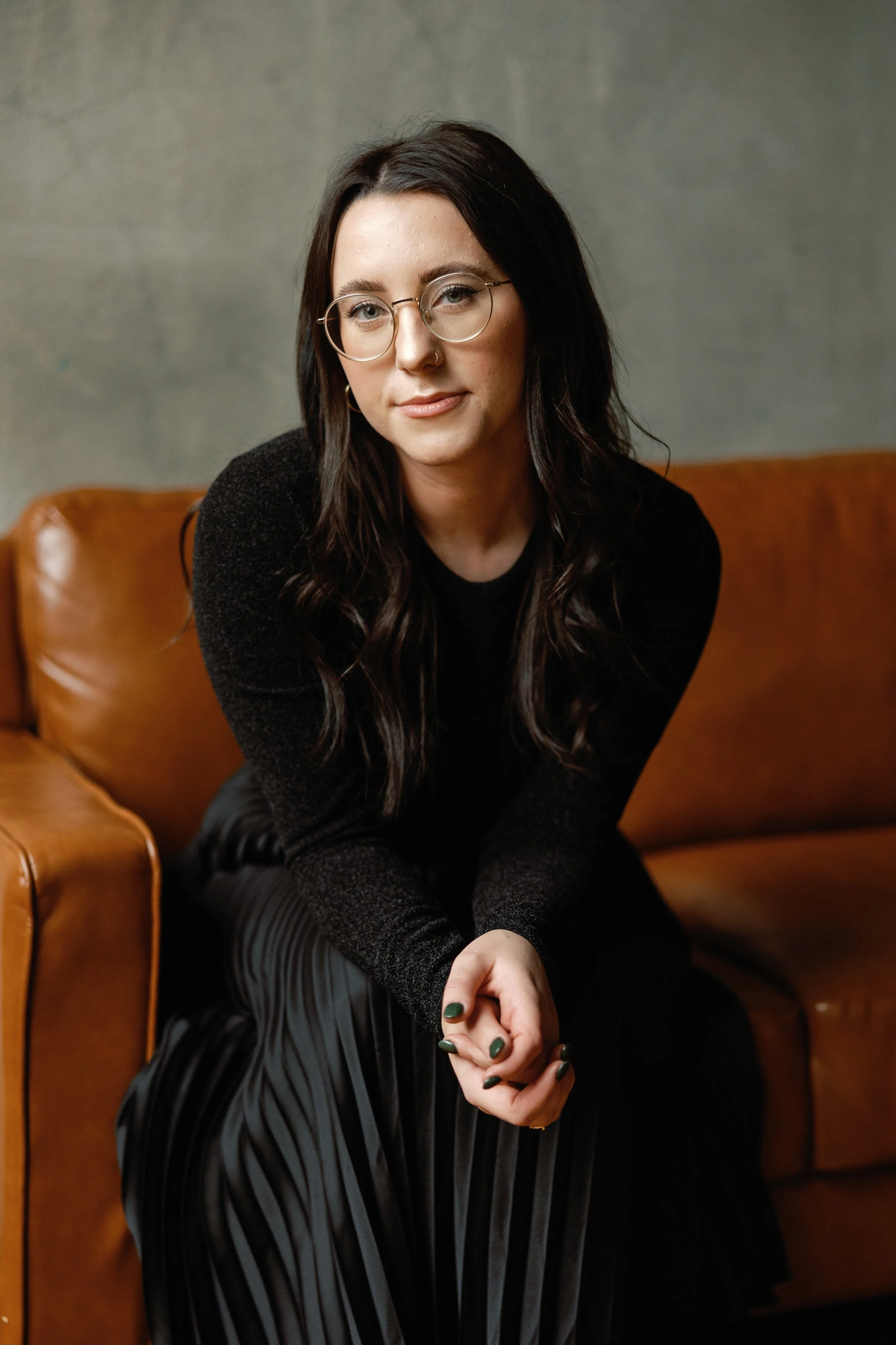 A Tennessee Photographer with long dark hair, wearing glasses, a nose ring, and a black sweater, sitting on a brown leather couch against a gray wall, with her hands clasped and looking at the camera.