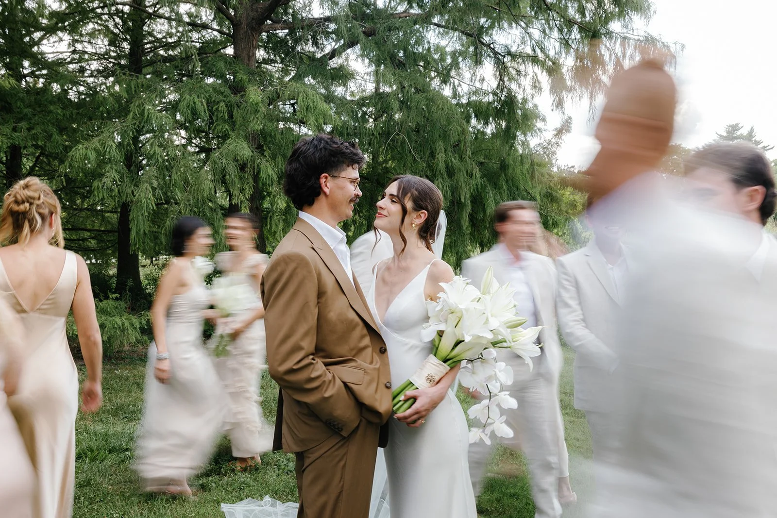 A bride and groom are smiling and gazing at each other at their outdoor wedding, with guests dancing and mingling in the background.