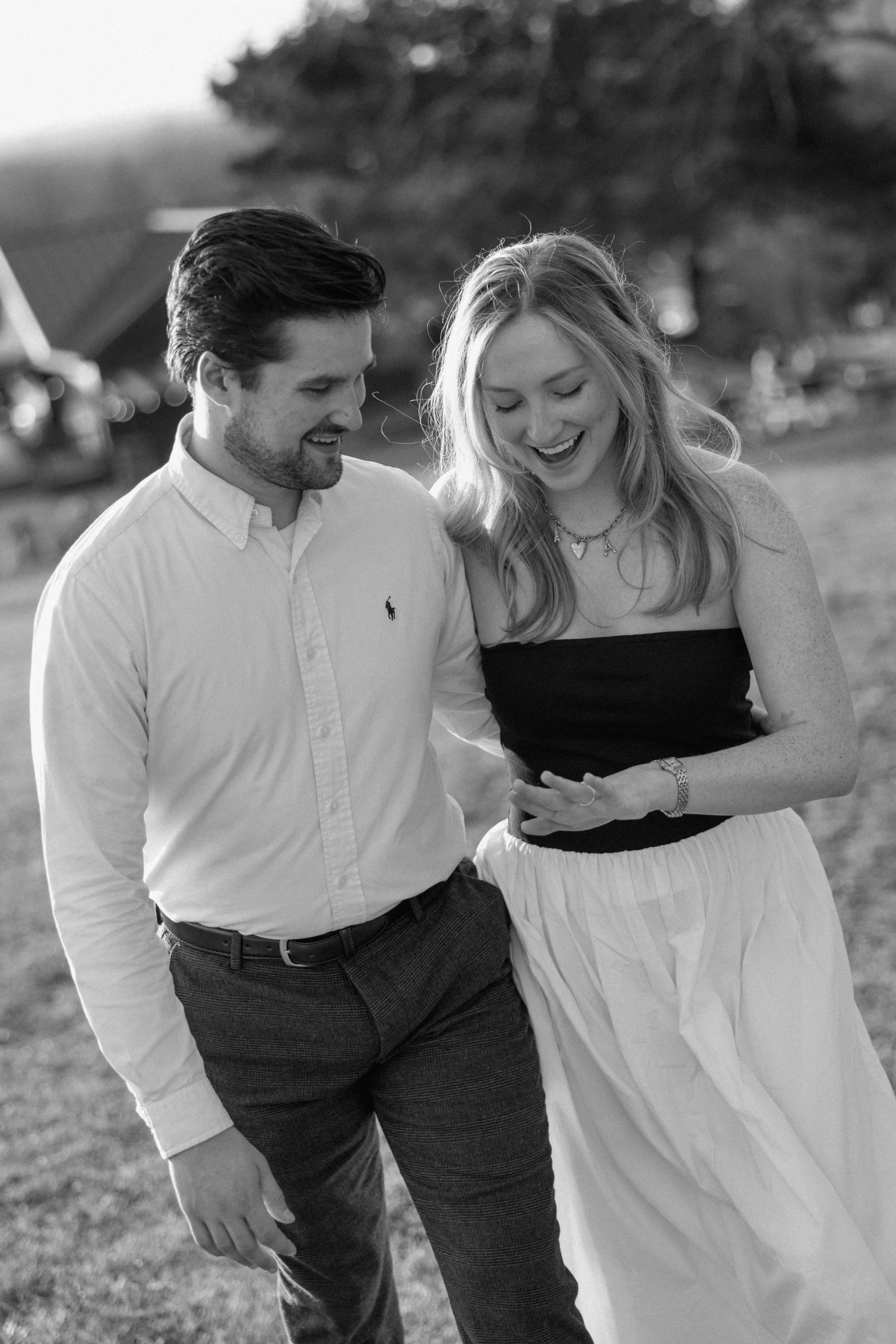 A young couple is walking outdoors smiling and looking at the engagement ring together, with a blurred background of trees in Tennessee.