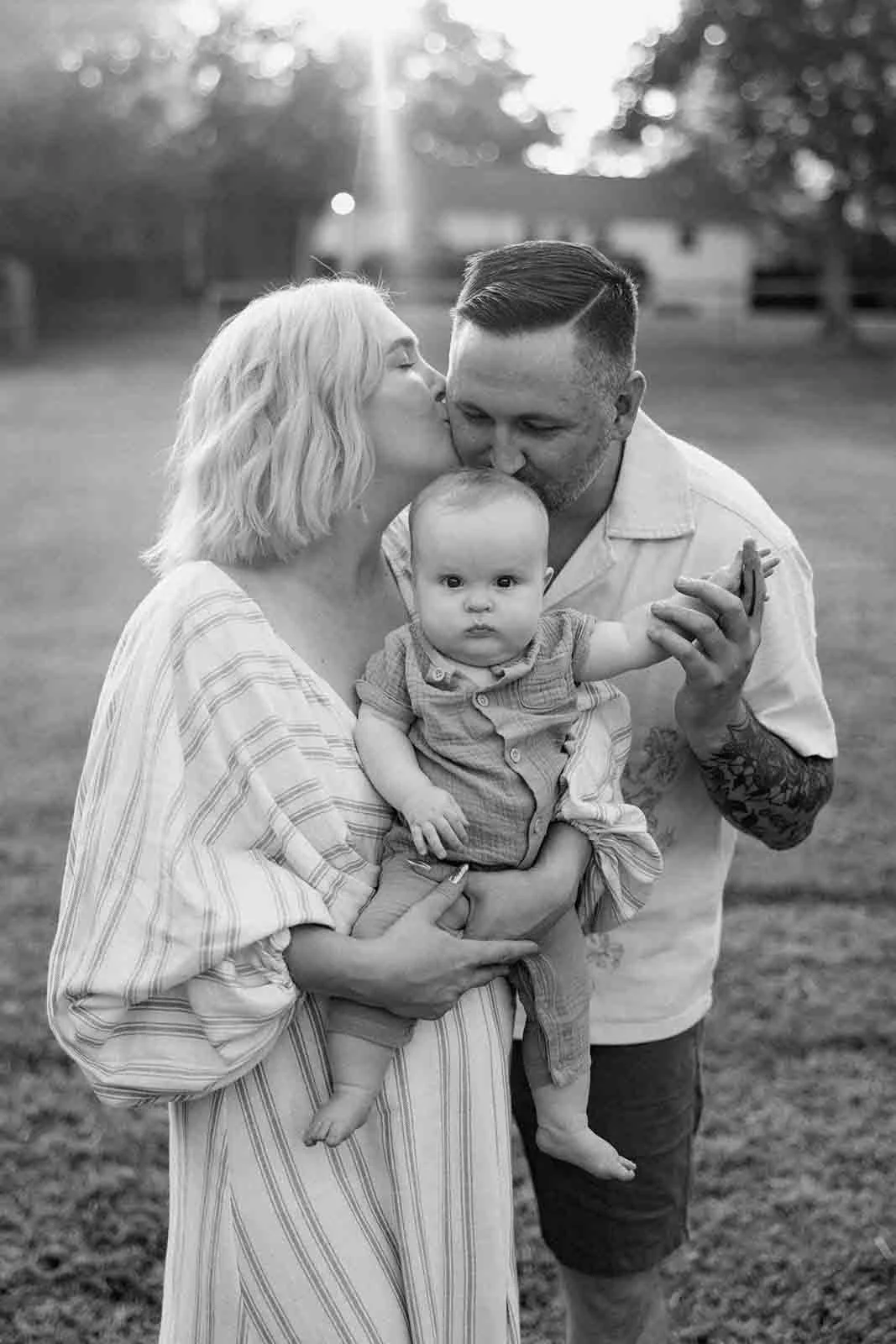 A black and white photo of a family outdoors: a woman with shoulder-length wavy hair holding a baby, a man with short hair kissing the baby's head