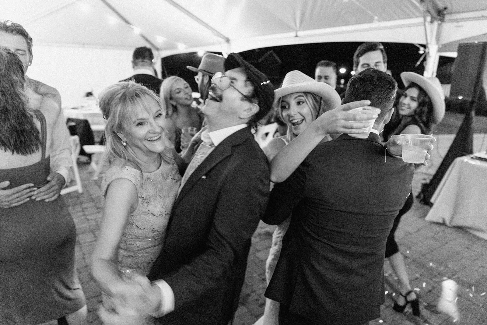 People dancing and celebrating at an outdoor party under a canopy, smiling and holding drinks, in a black and white photo.