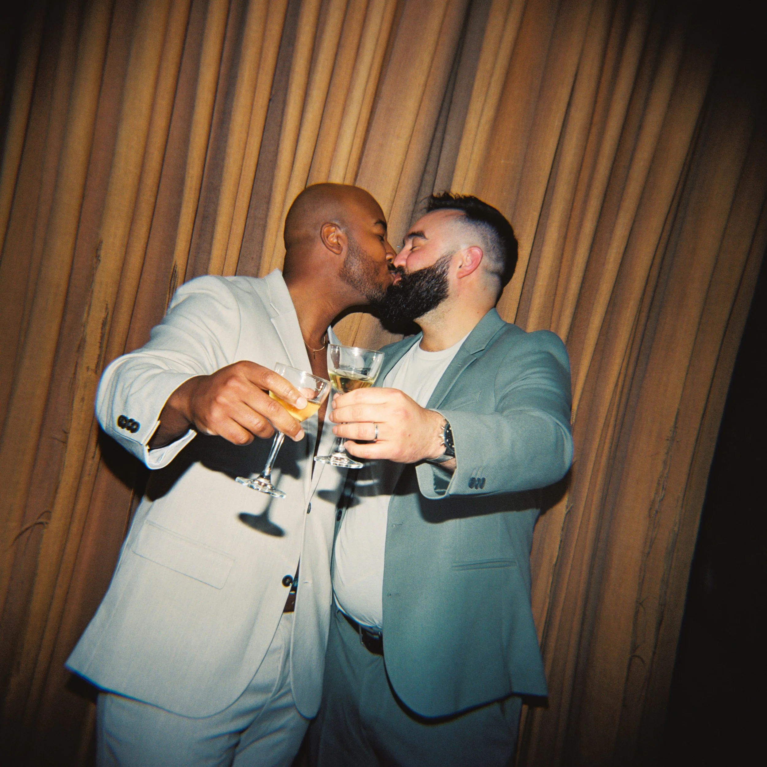 Two men dressed in nice suits holding glasses out and kissing with a mustard curtain background