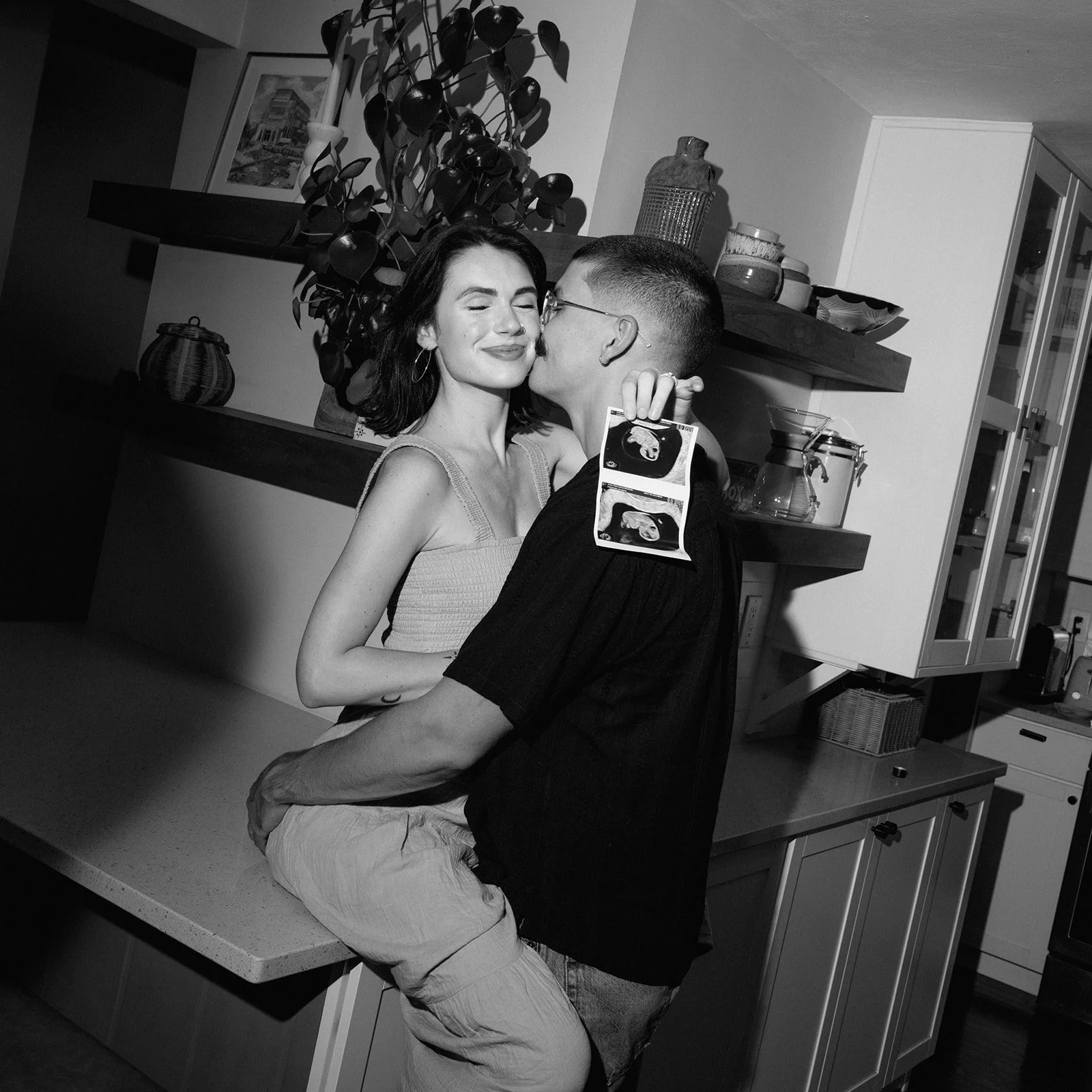 Black and white photo of a woman sitting on a counter hugging her husband while holding ultrasound photos