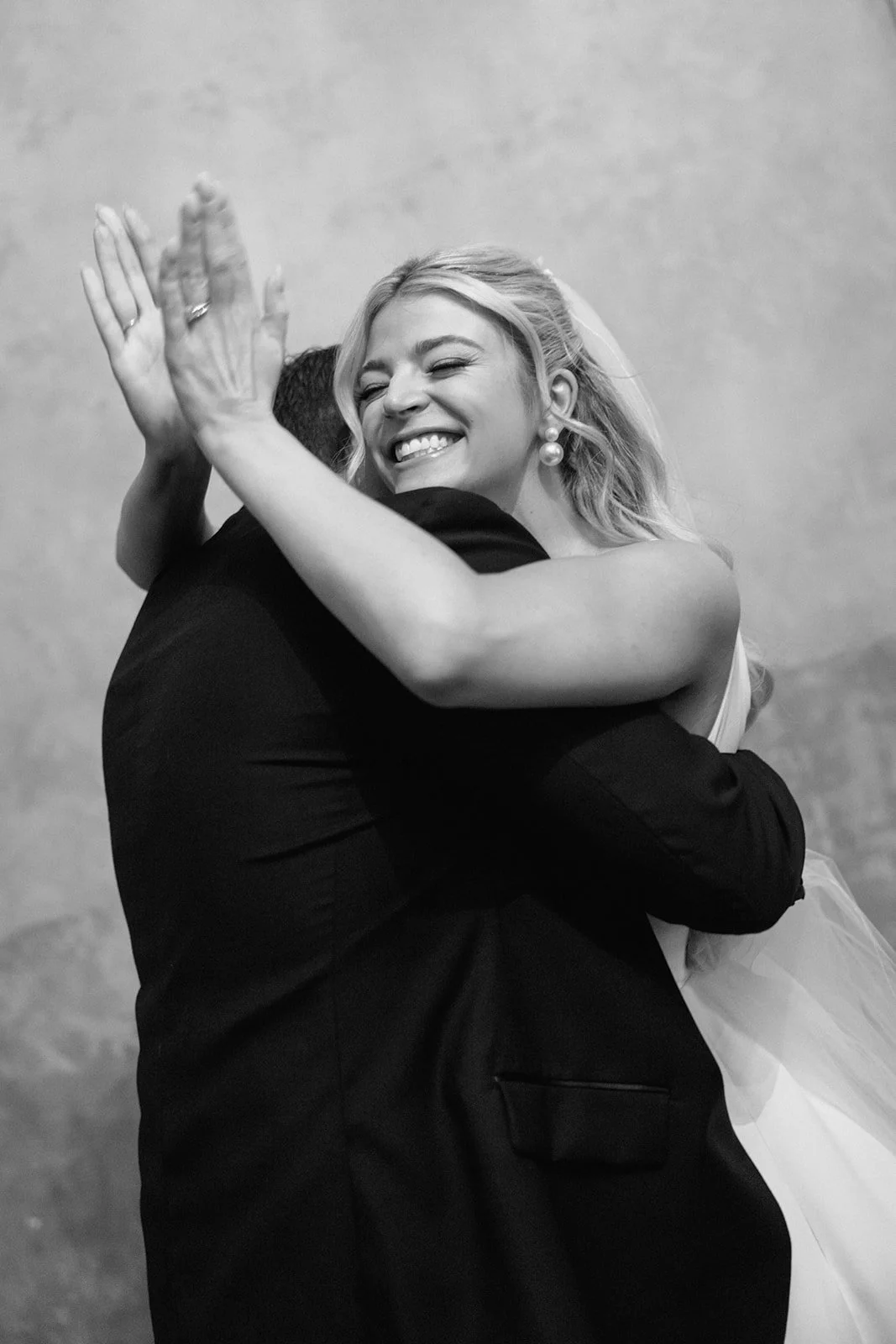 Black and white photo of a bride hugging a person in a dark suit, smiling with her eyes closed.
