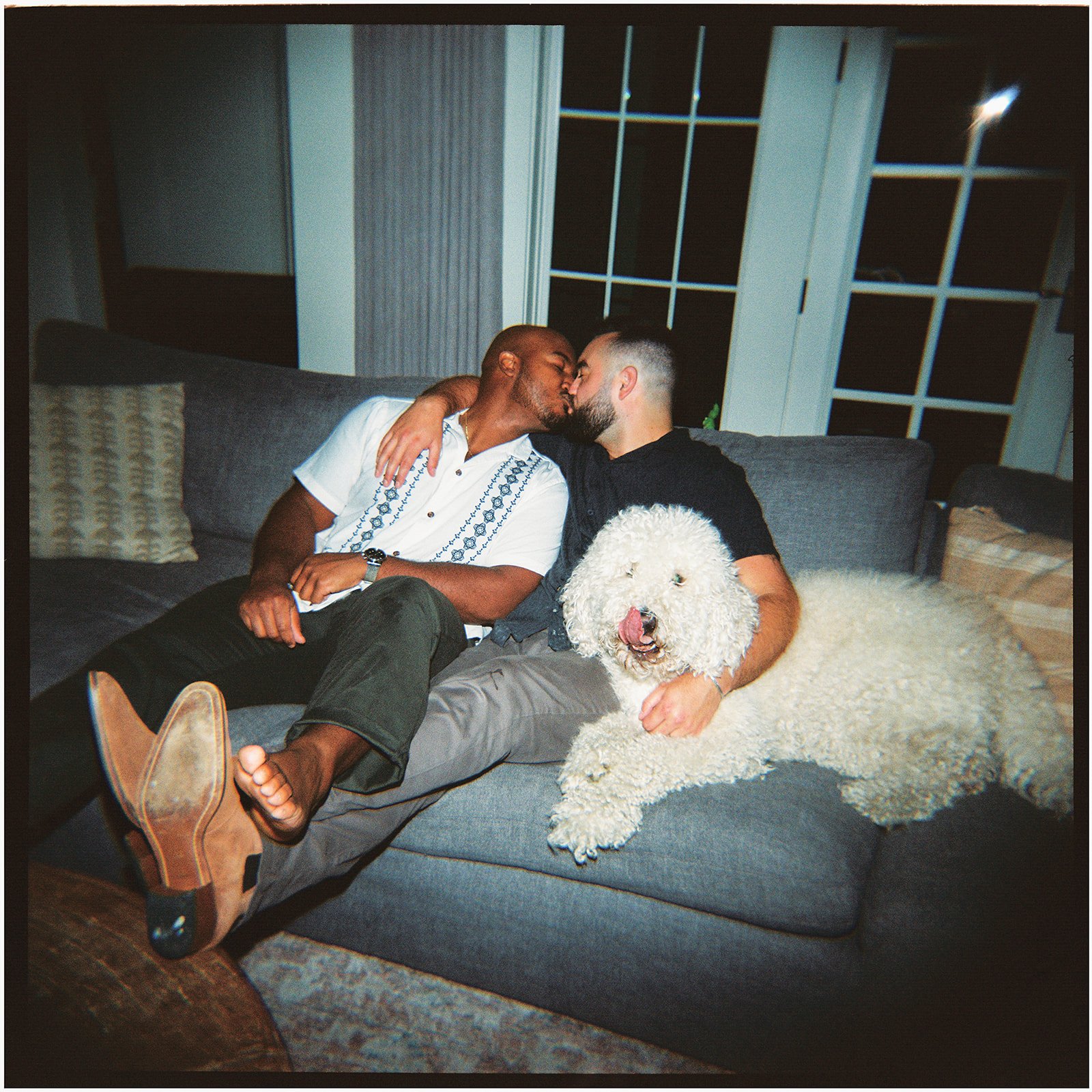 Two men kissing on a couch in the living room with their dog