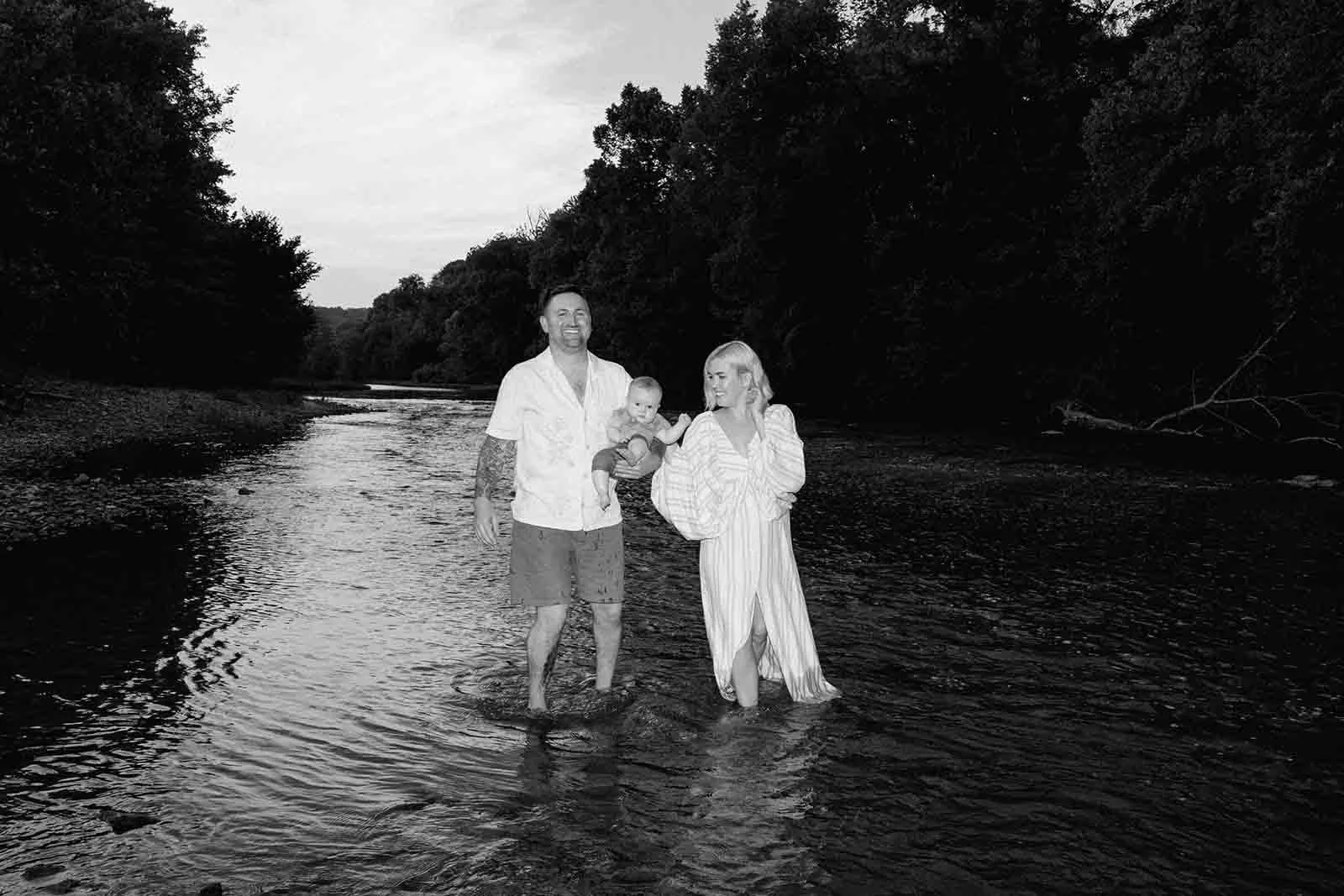 Black and white photo of a family of three standing in a stream in matching cream attire