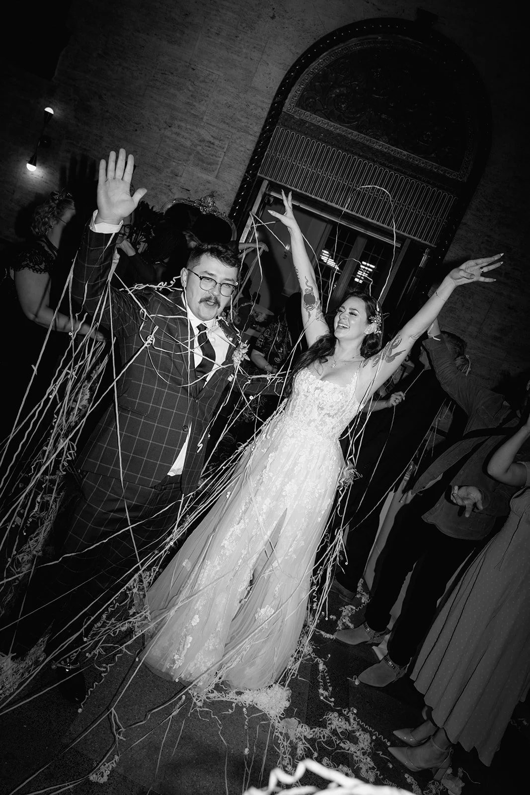 A newlywed couple celebrating with friends at their wedding reception, surrounded by streamers and confetti. The bride is wearing a wedding dress, while the groom is dressed in a suit.