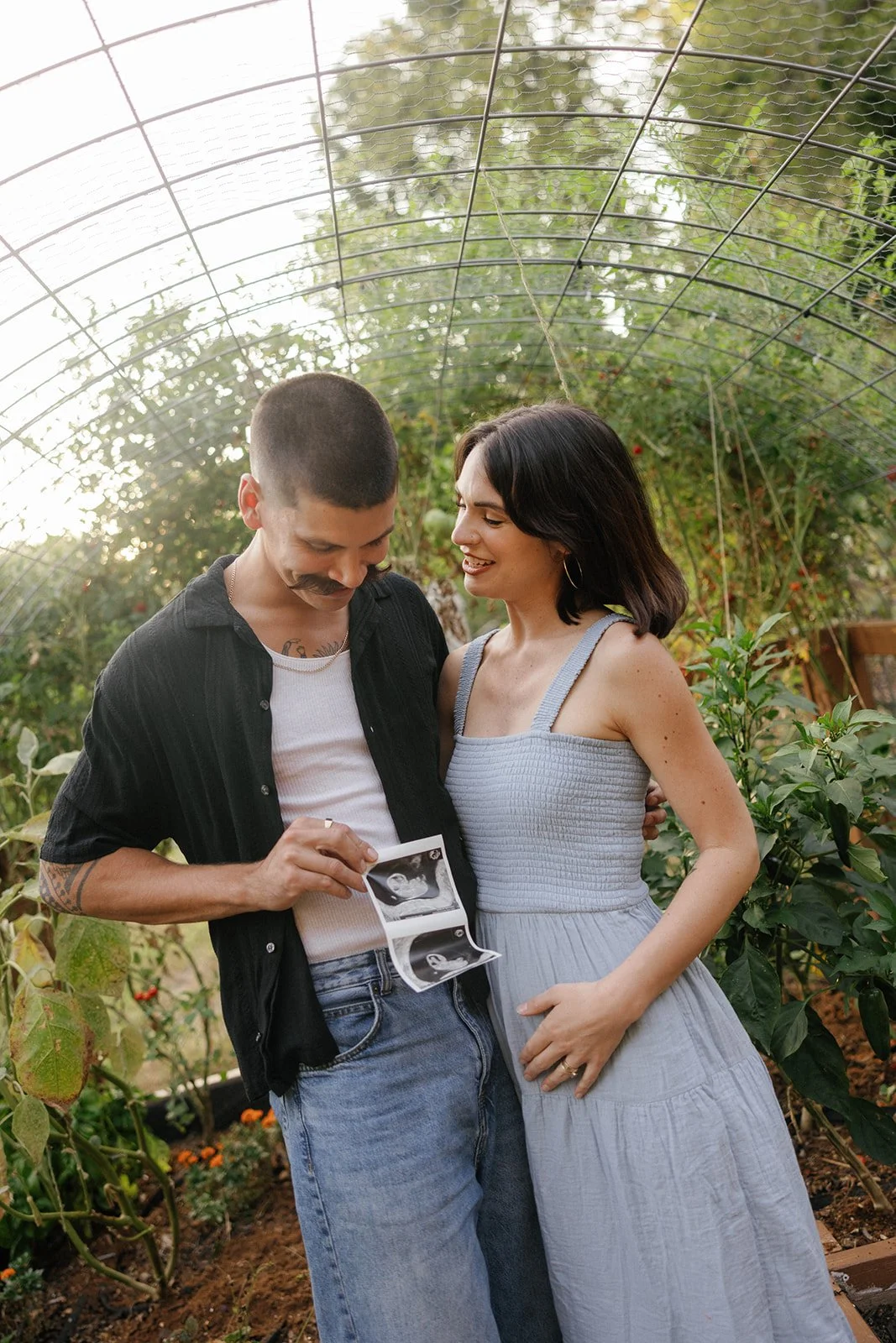 Husband holding ultrasound photos in a garden while standing next to his pregnant wife