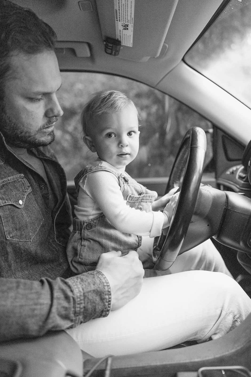 Black and white photo of a baby sitting on his dad's lap while holding the steering wheel