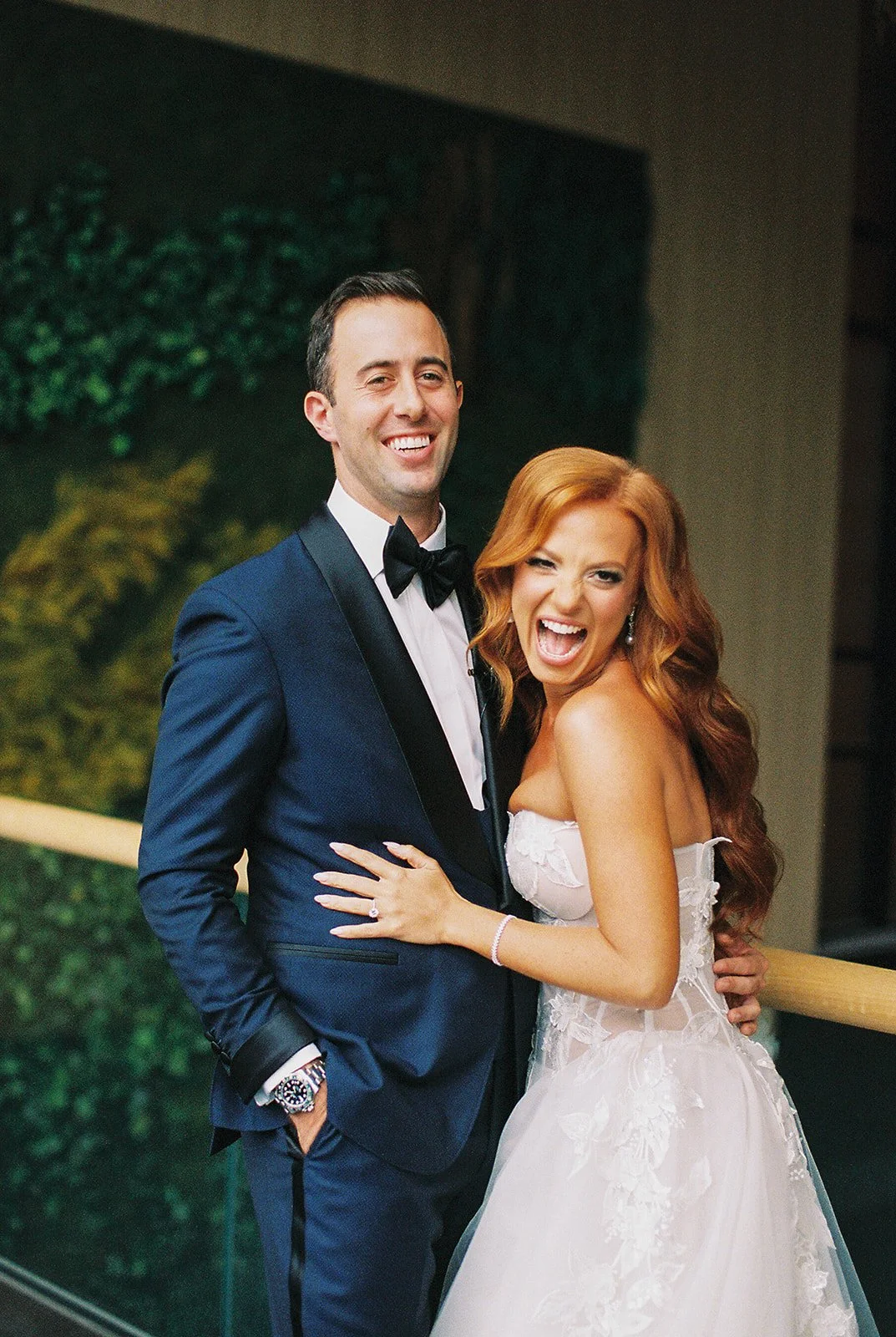 A smiling man in a dark blue tuxedo and bow tie standing next to a woman in a white wedding dress, both smiling and posing for a photo.