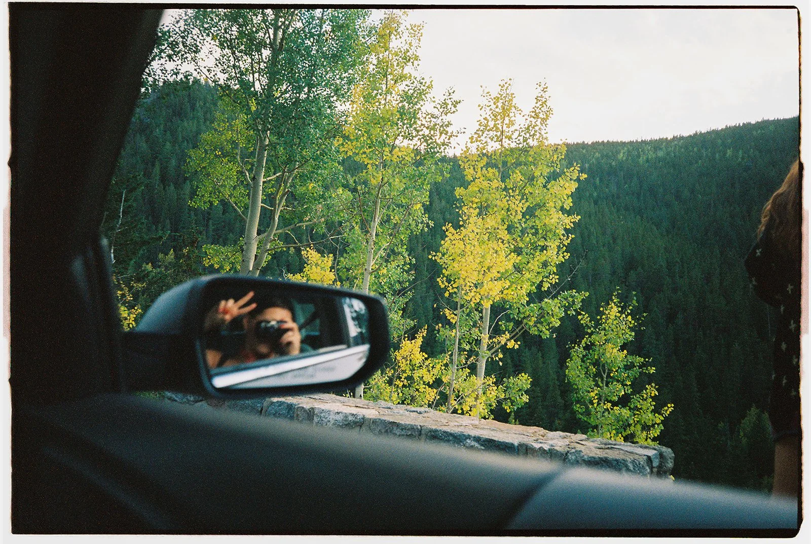 Photo taken from inside a vehicle showing the side mirror, with a person taking a photo. Outside the vehicle, there's a scenic view of green and yellow trees and a forested mountain landscape.