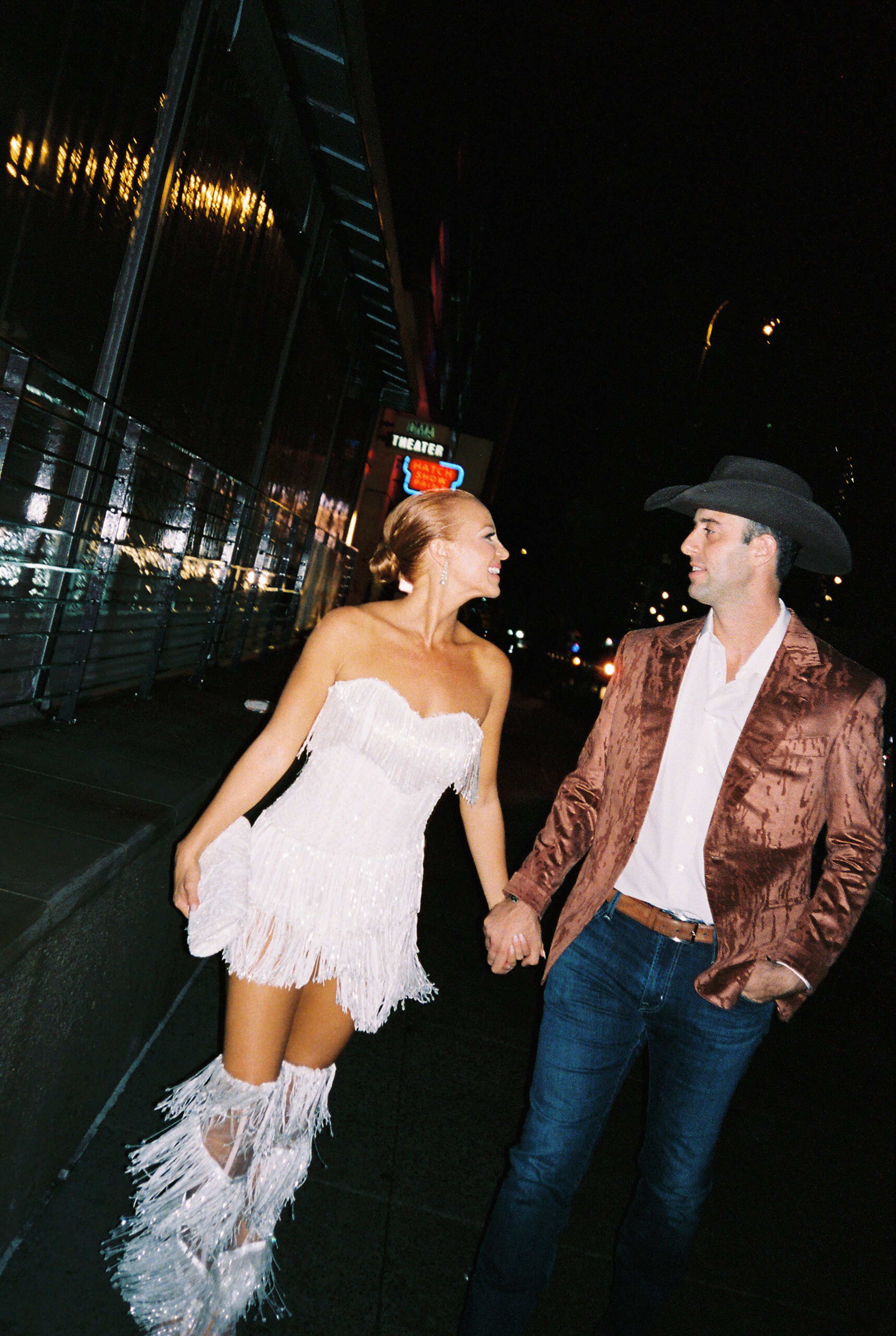A woman wearing a white, fringed strapless dress and matching shiny boots holding hands with a man in a brown textured blazer, white shirt, and a cowboy hat, standing on a city street at night.