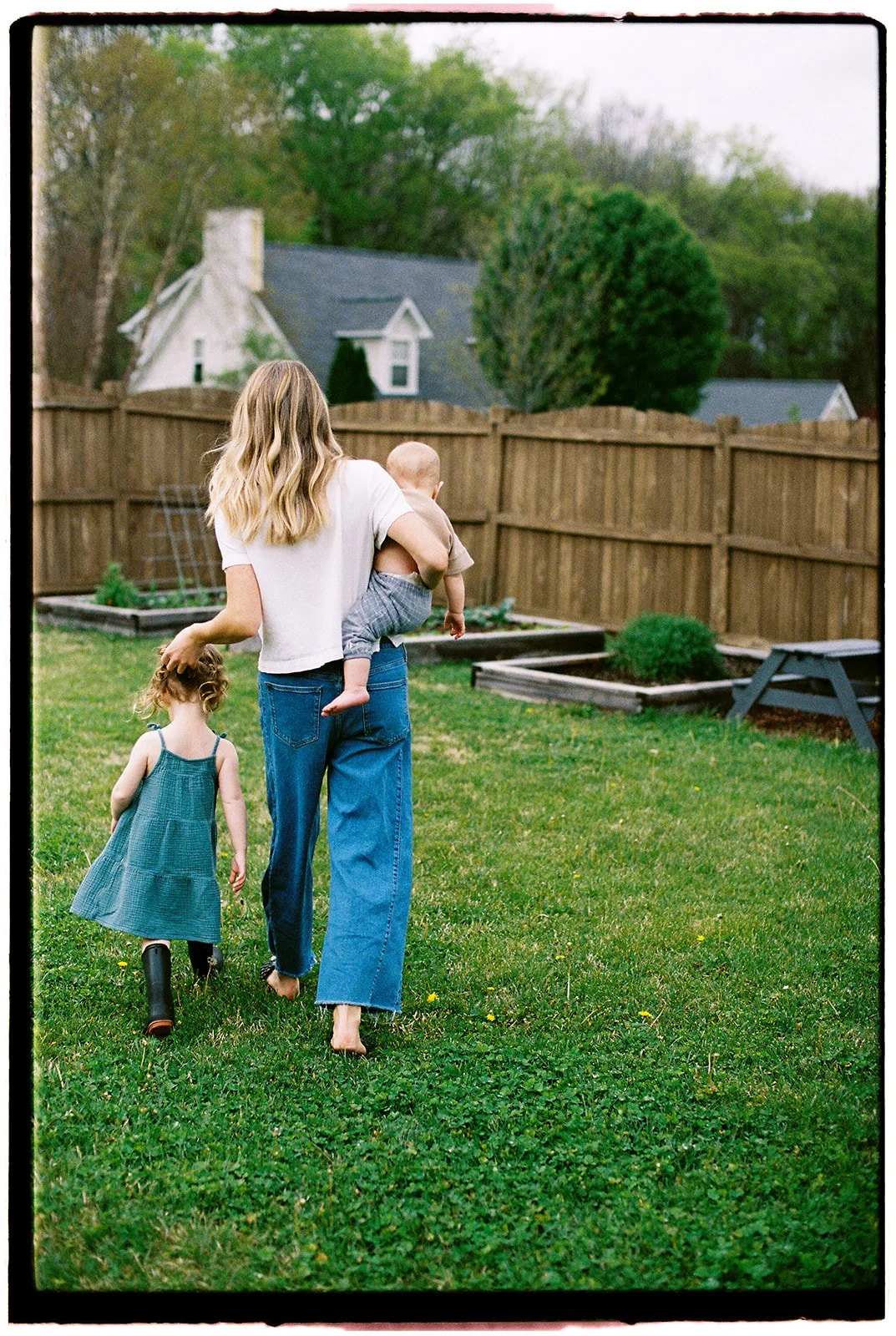 Film photo of a woman walking with a baby on her hip and holding the top of her other child's head while walking to their backyard garden