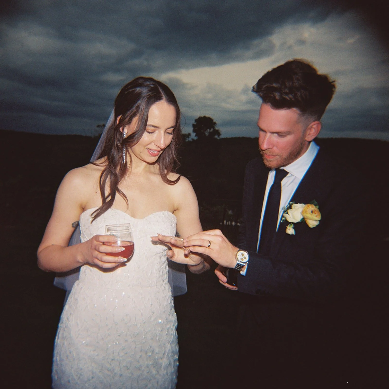 A film photo of a bride and groom exchanging rings outdoors during a wedding ceremony at dusk.