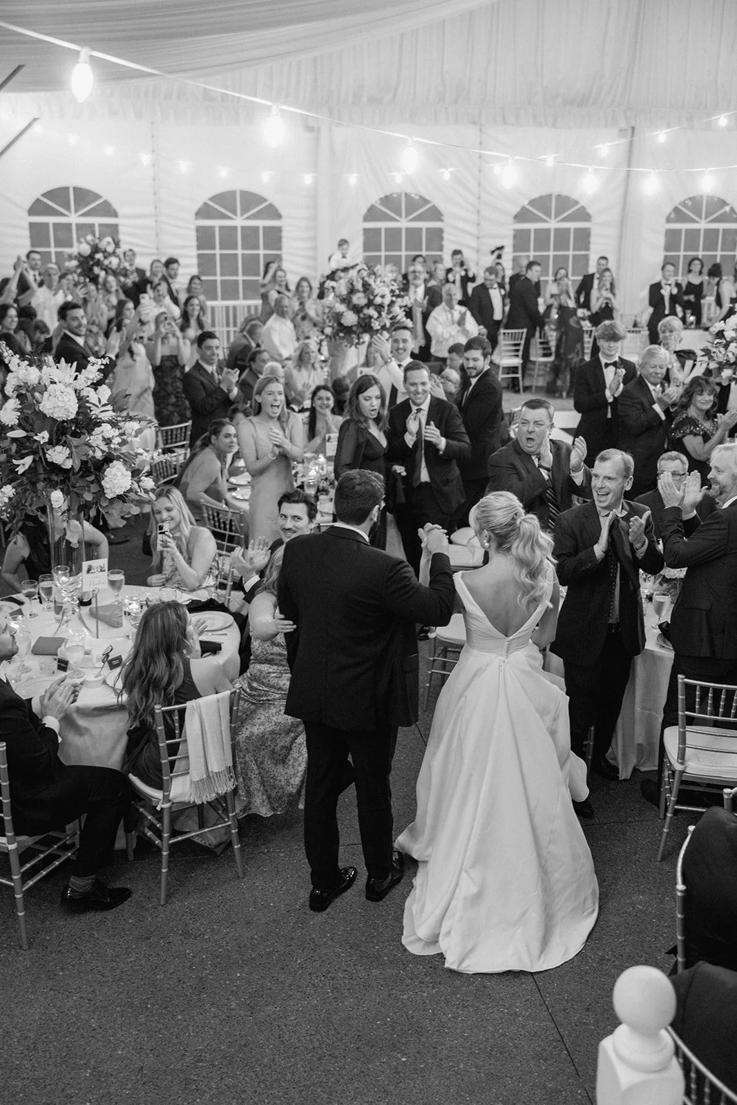 Black and white photo of a wedding reception with bride and groom celebrate in front of a large crowd at decorated tables.