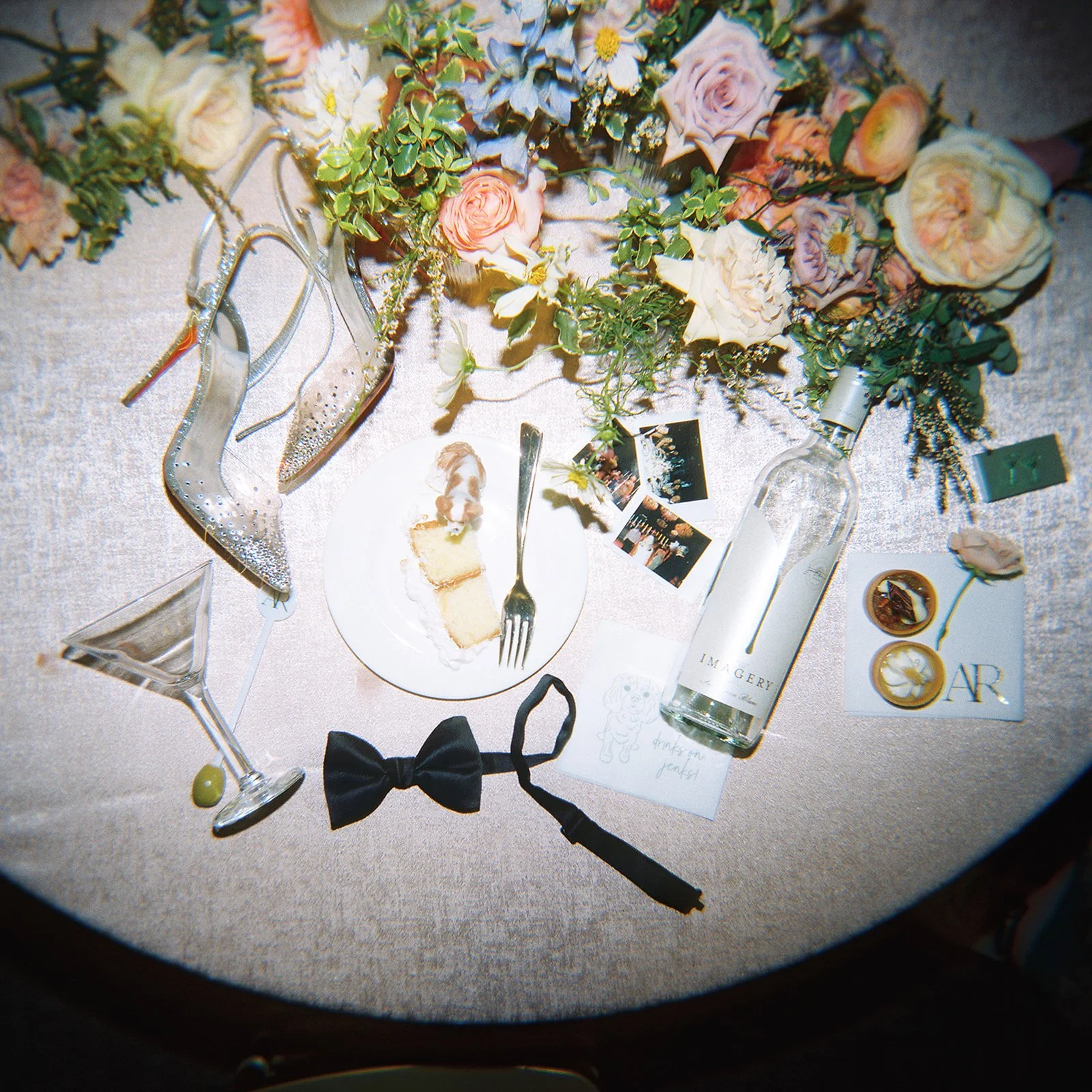 A table decorated with pink and white flowers and greenery, including roses and hydrangeas. On the table, there are a pair of silver high-heeled shoes, a pair of sunglasses, a small portion of cake on a plate with a fork, some photographs, a bottle of vodka, a black bowtie, a paper with handwritten notes, and a card with the words 'Thank you guests!'