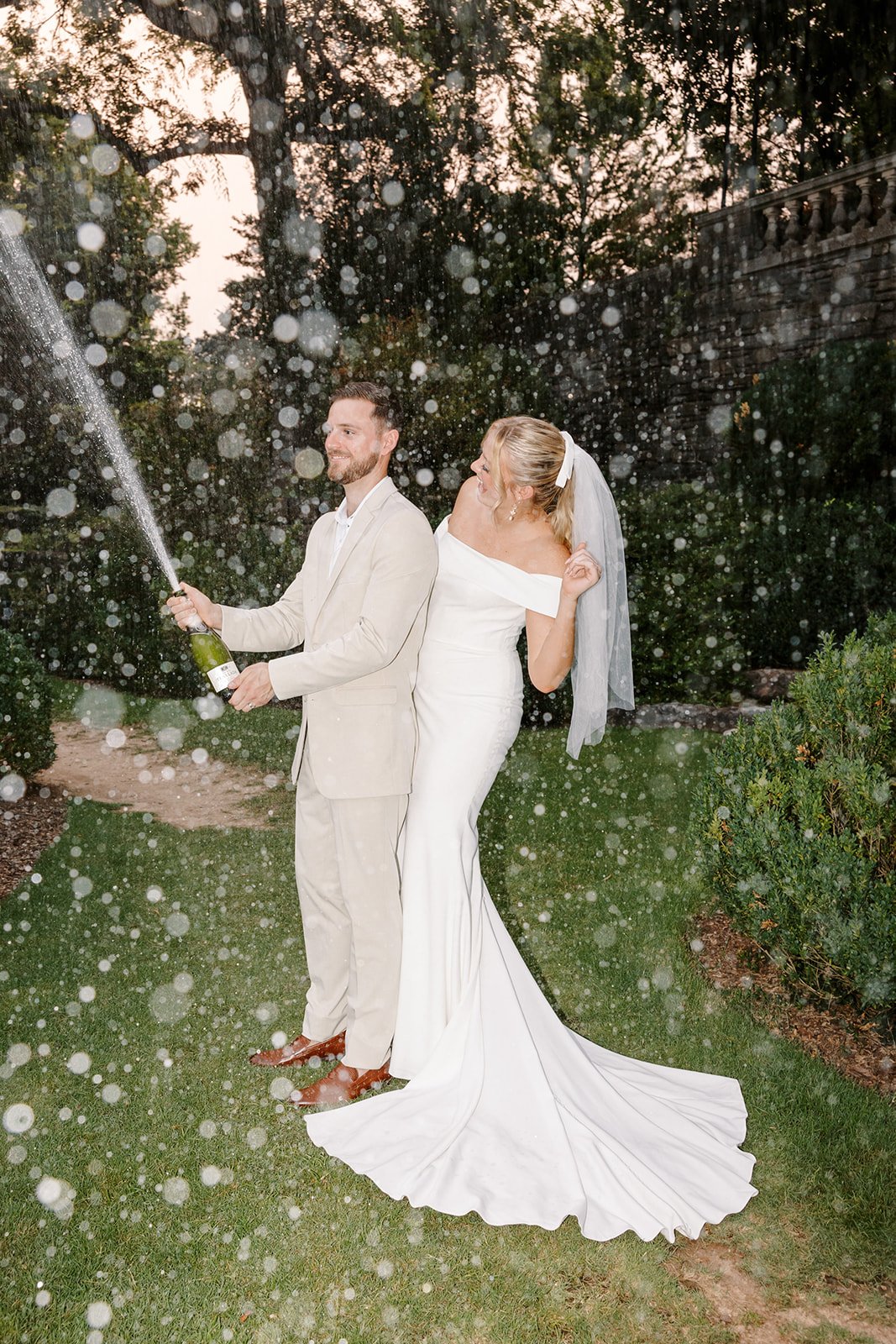 Bride and groom celebrating outdoors, with the groom opening a champagne bottle, causing a spray of champagne, during their wedding.
