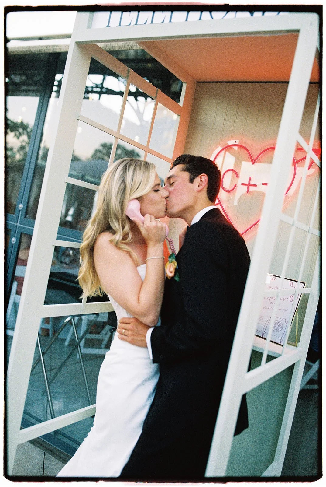 A bride and groom sharing a kiss at their wedding, with a neon heart in the background.