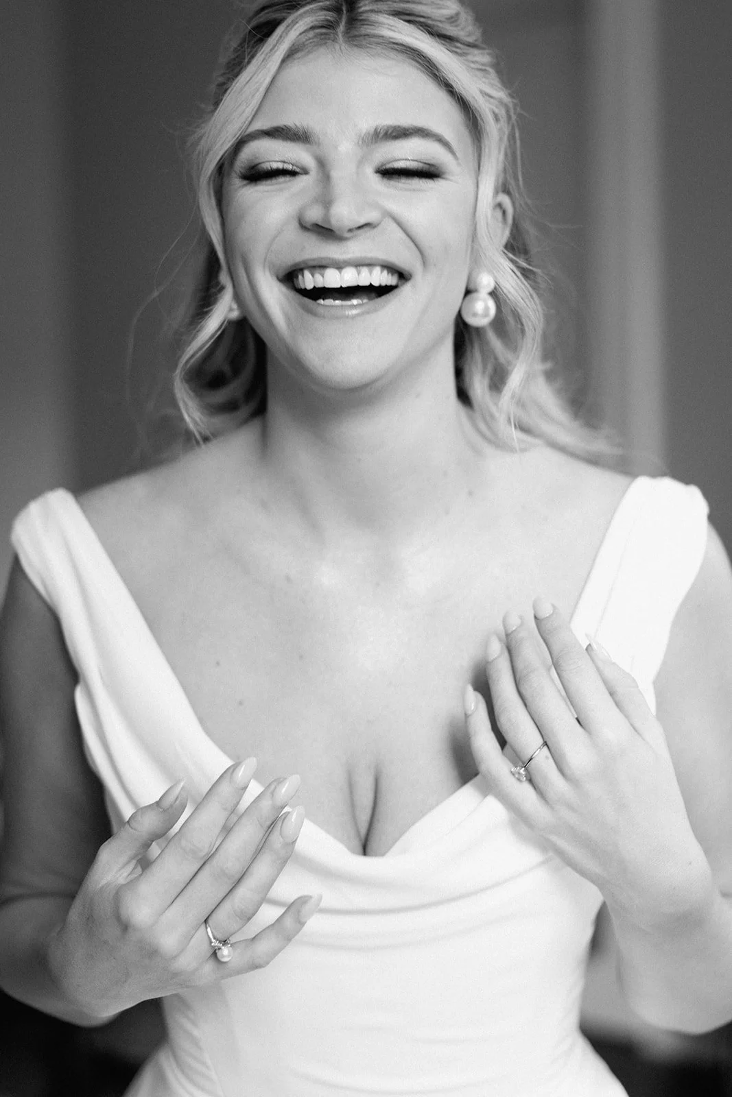 A smiling woman in a white wedding dress with earrings, holding her hands over her chest, in a black and white photo.