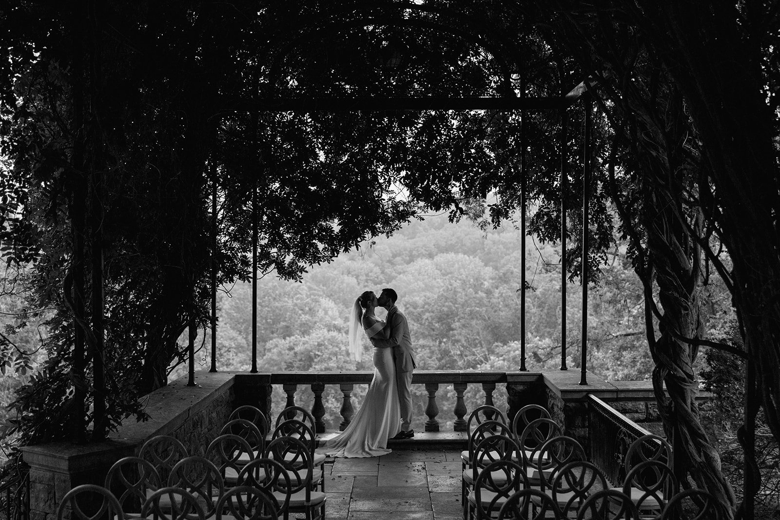 A black-and-white photo of a bride and groom sharing a kiss on a small stone outlook, surrounded by trees and overlooking a forested landscape at Cheekwood Gardens.
