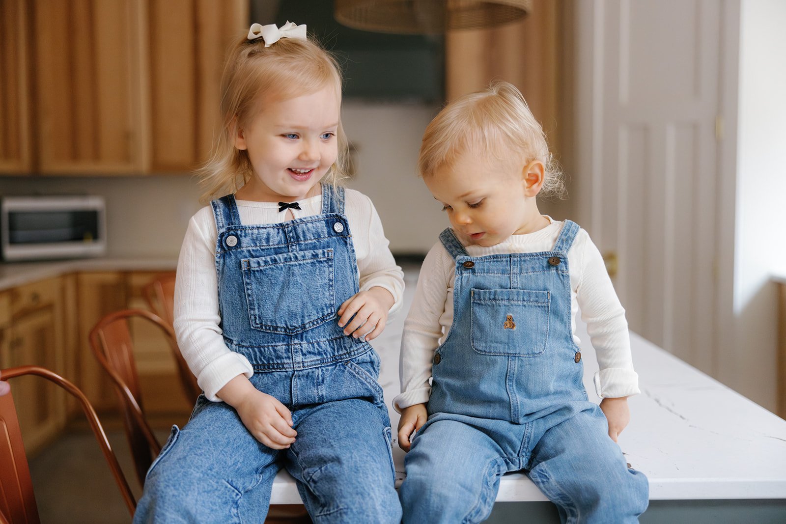 Siblings in matching overall outfits sit on the counter of their home playing and smiling