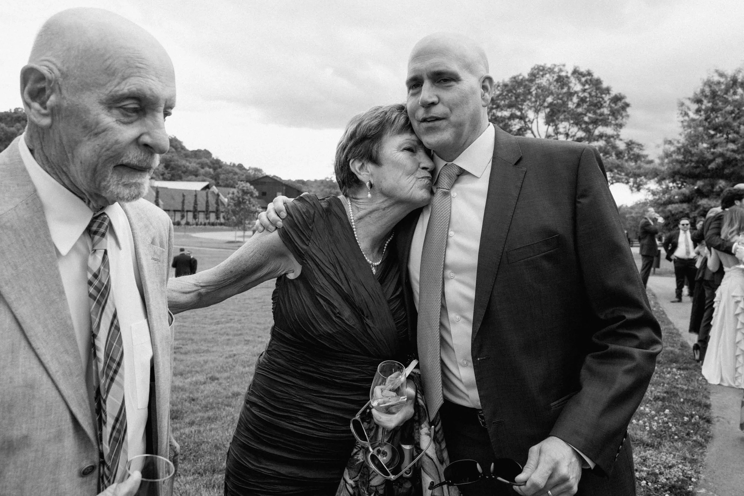 Black and white photo of a woman holding on to an older man's arm while leaning into hug another man at a wedding while they are all formally dressed