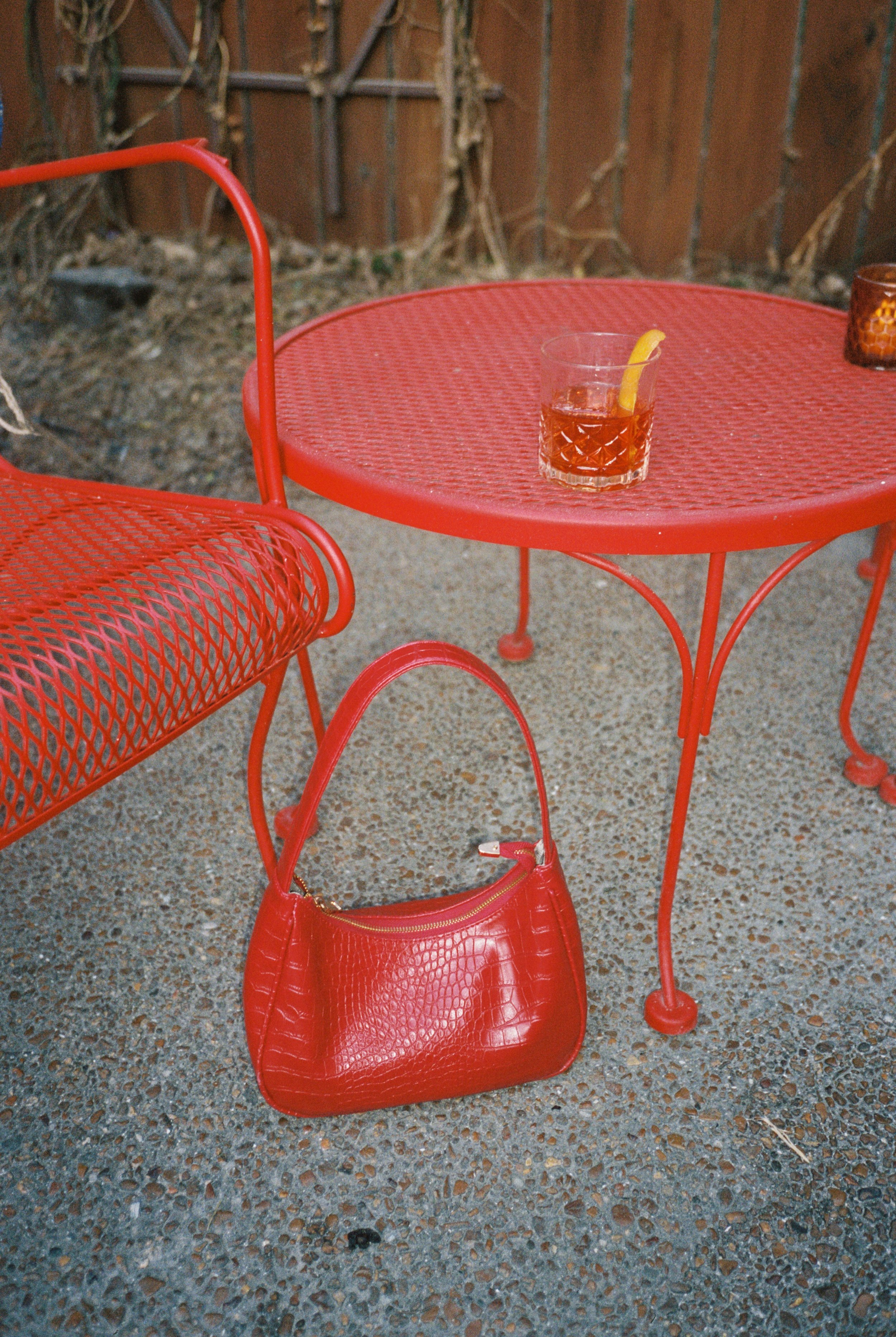 Red metal outdoor table with a glass of dark beverage and a lemon wedge, red metal chair, red handbag on the ground, brown wooden fence and dried plants in background, gravel ground.