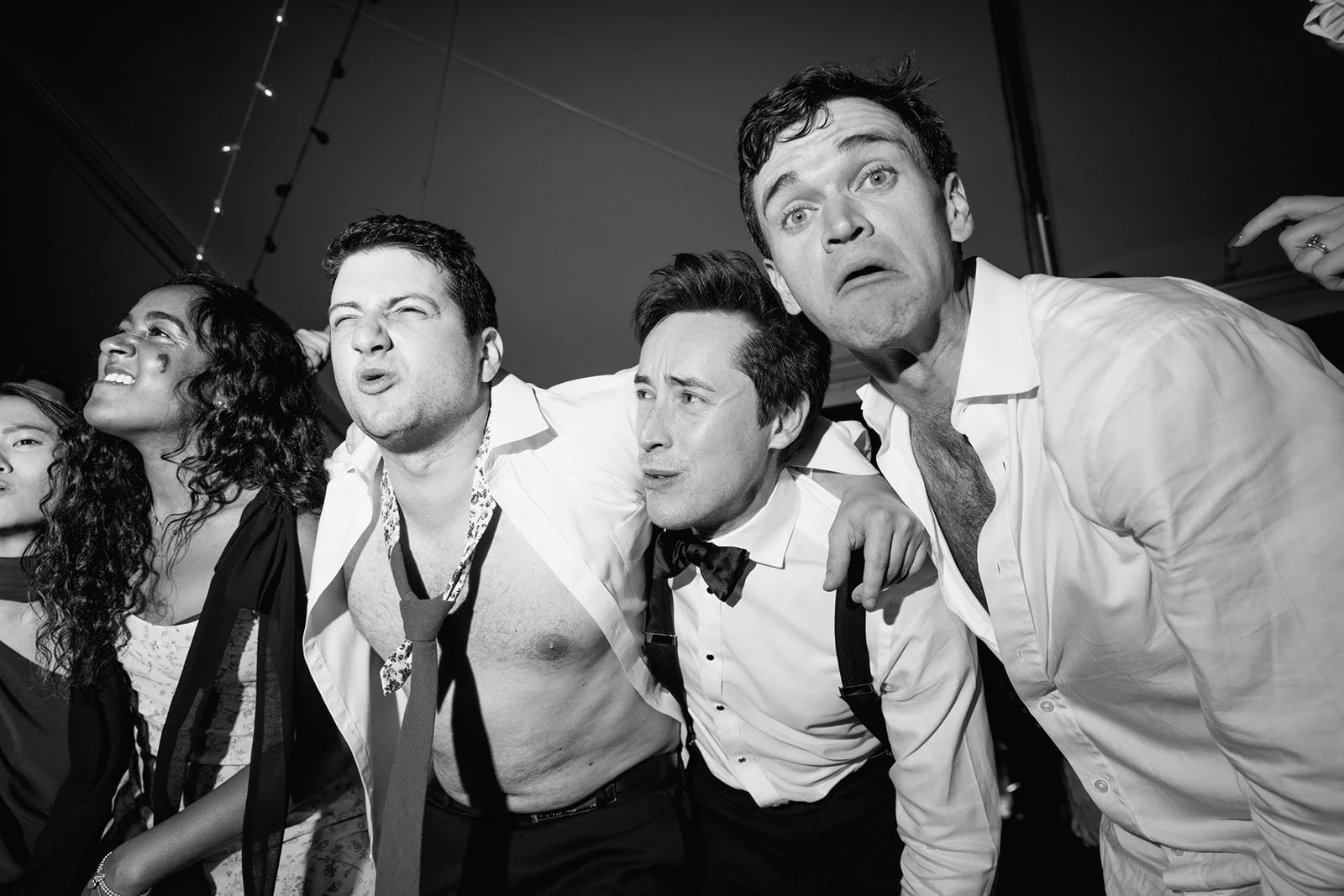 Black and white photo of five people standing closely with arms around each other's shoulders, making exaggerated facial expressions, at a wedding.